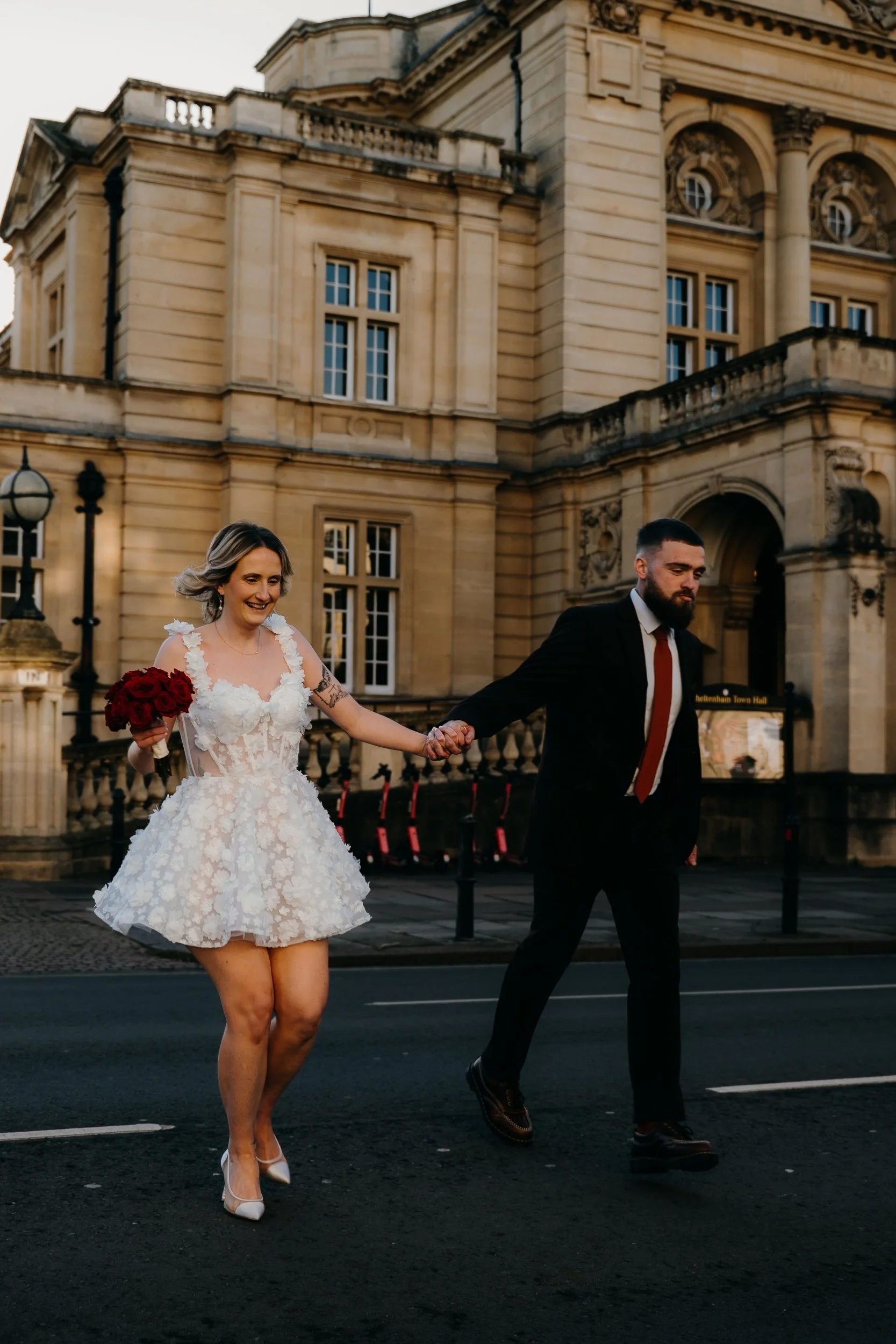 A woman in a white lace dress holding a bouquet of red roses, smiling as she walks hand-in-hand with a man dressed in a black suit, on a city street in front of a historic building.