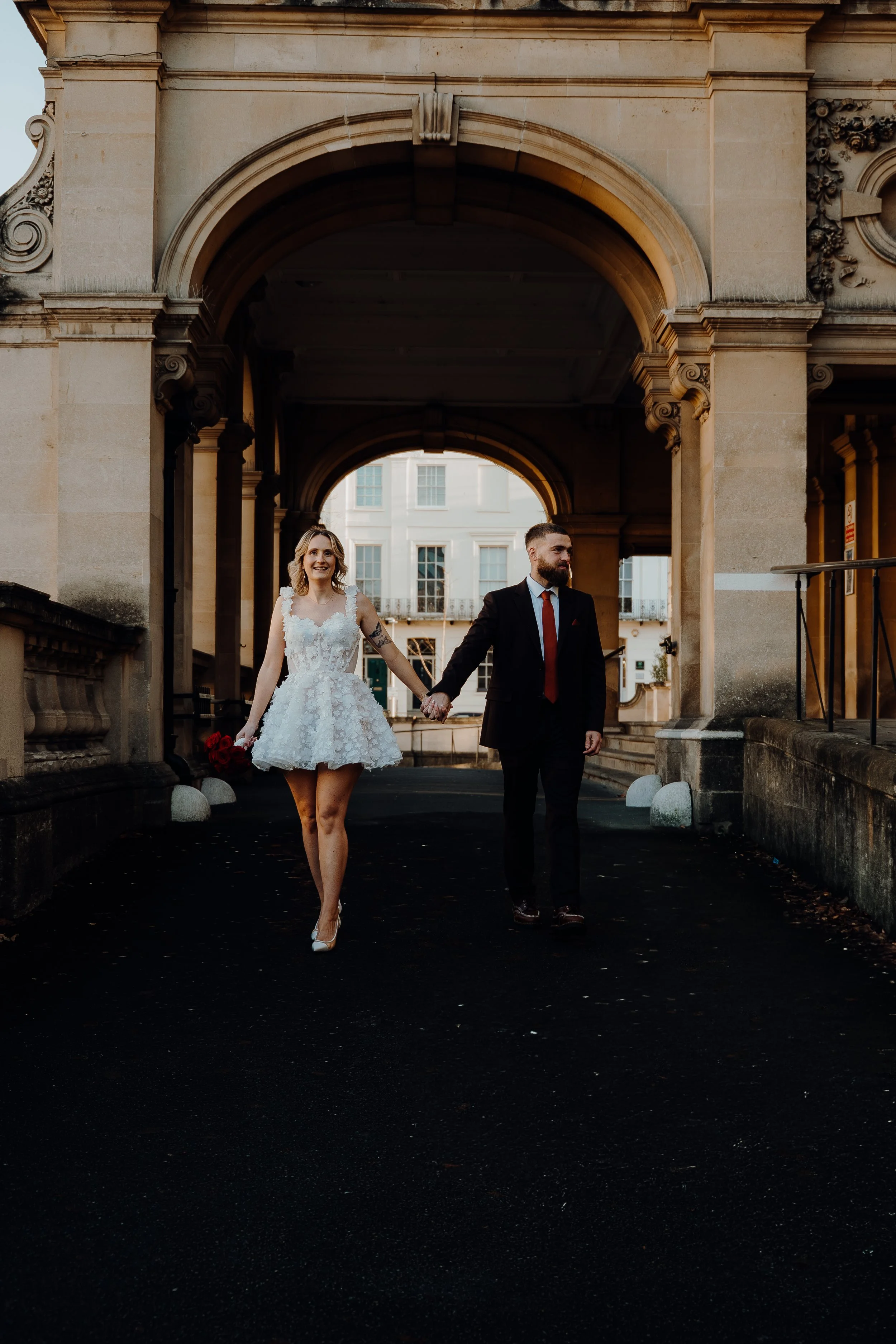 A smiling woman in a white lace dress holding a bouquet of red roses walking hand-in-hand with a man in a black suit, white shirt, and red tie under an ornate stone archway.