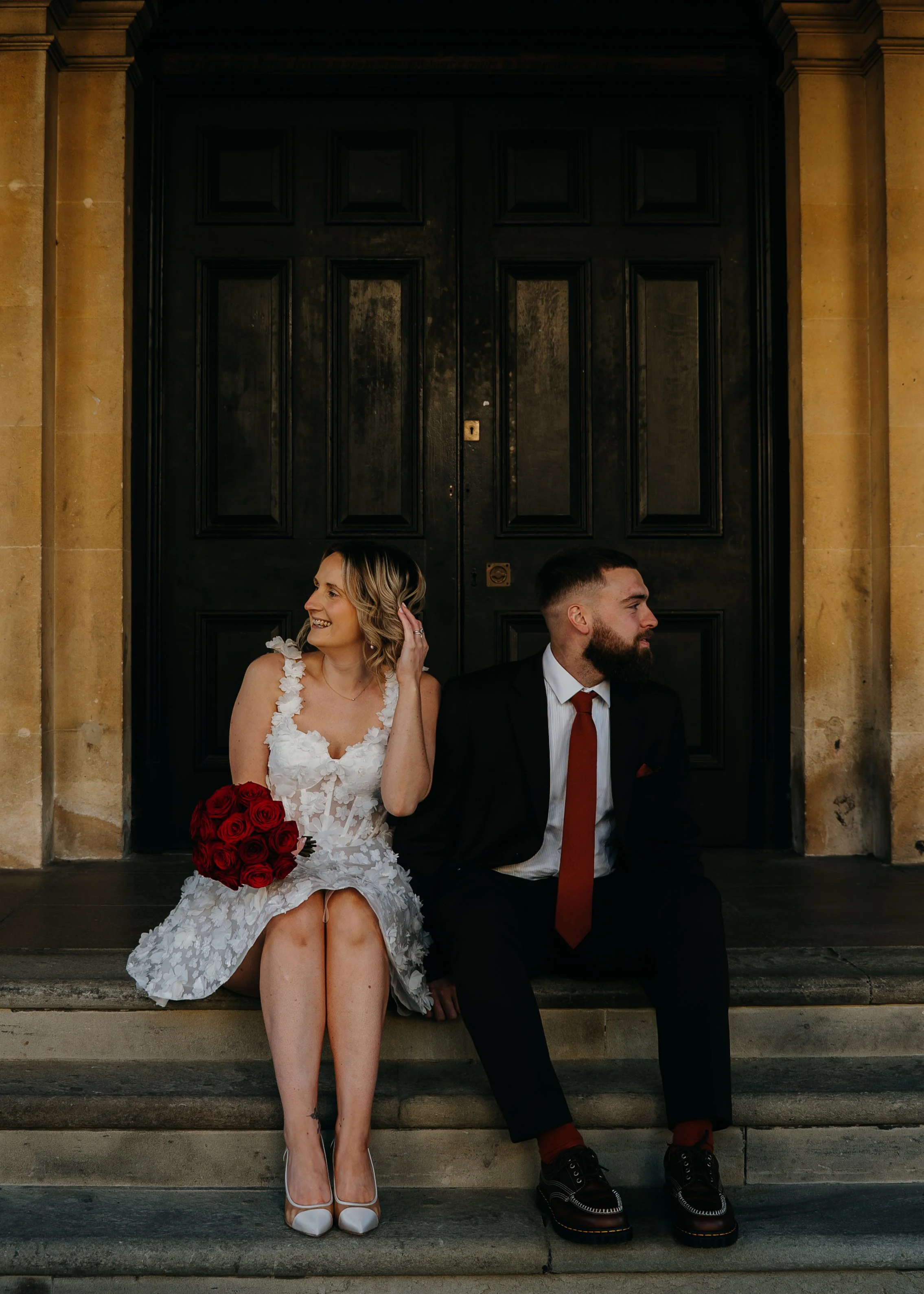 A bride and groom sitting on the steps in front of a large black wooden door, smiling and looking at each other. Iris willow photography bar 131 Cheltenham