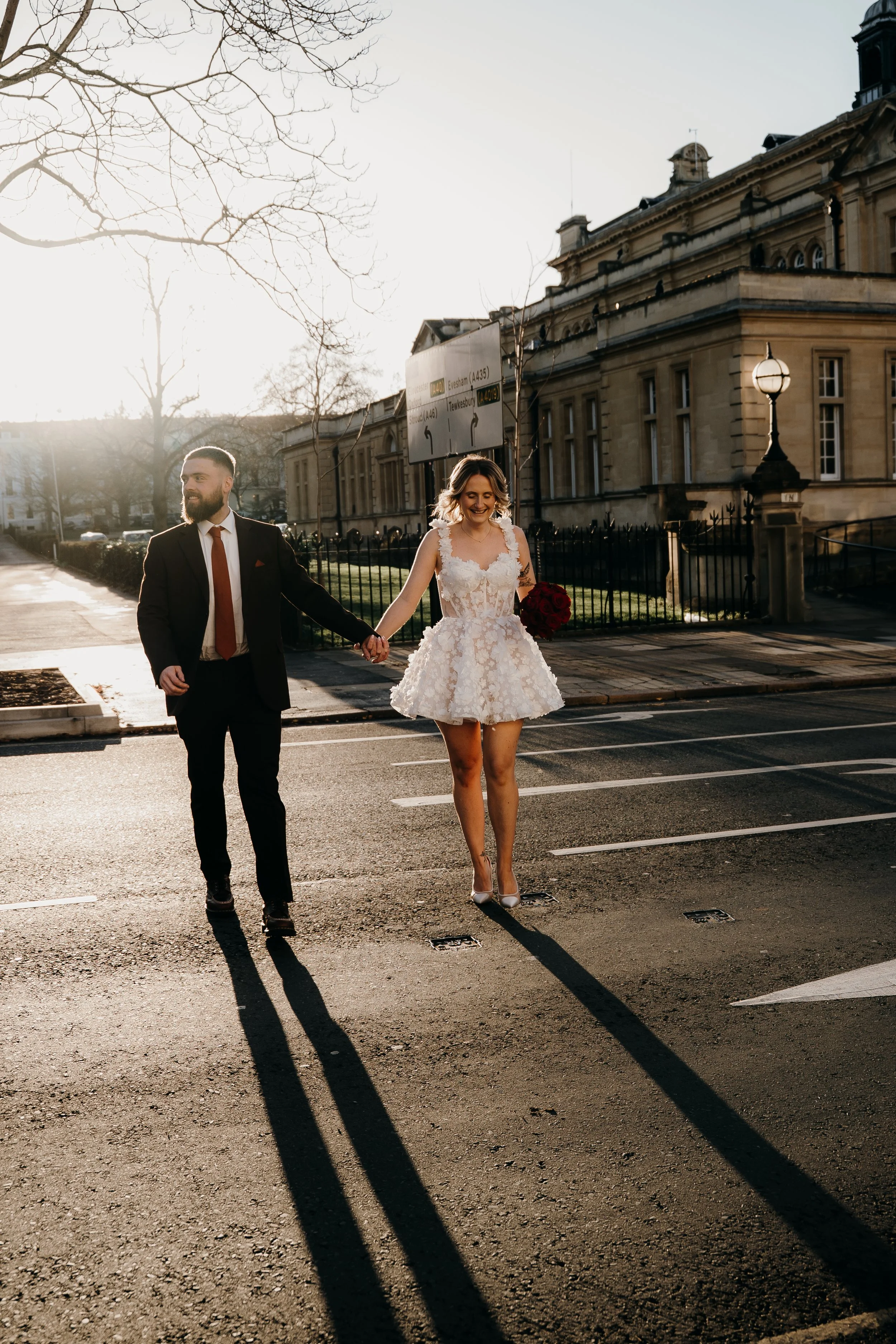 A man and woman walk hand in hand across a street during sunset. The woman is in a white dress holding a bouquet, and the man in a dark suit with a red tie. They are smiling, and the shadow of their figures stretches across the pavement.