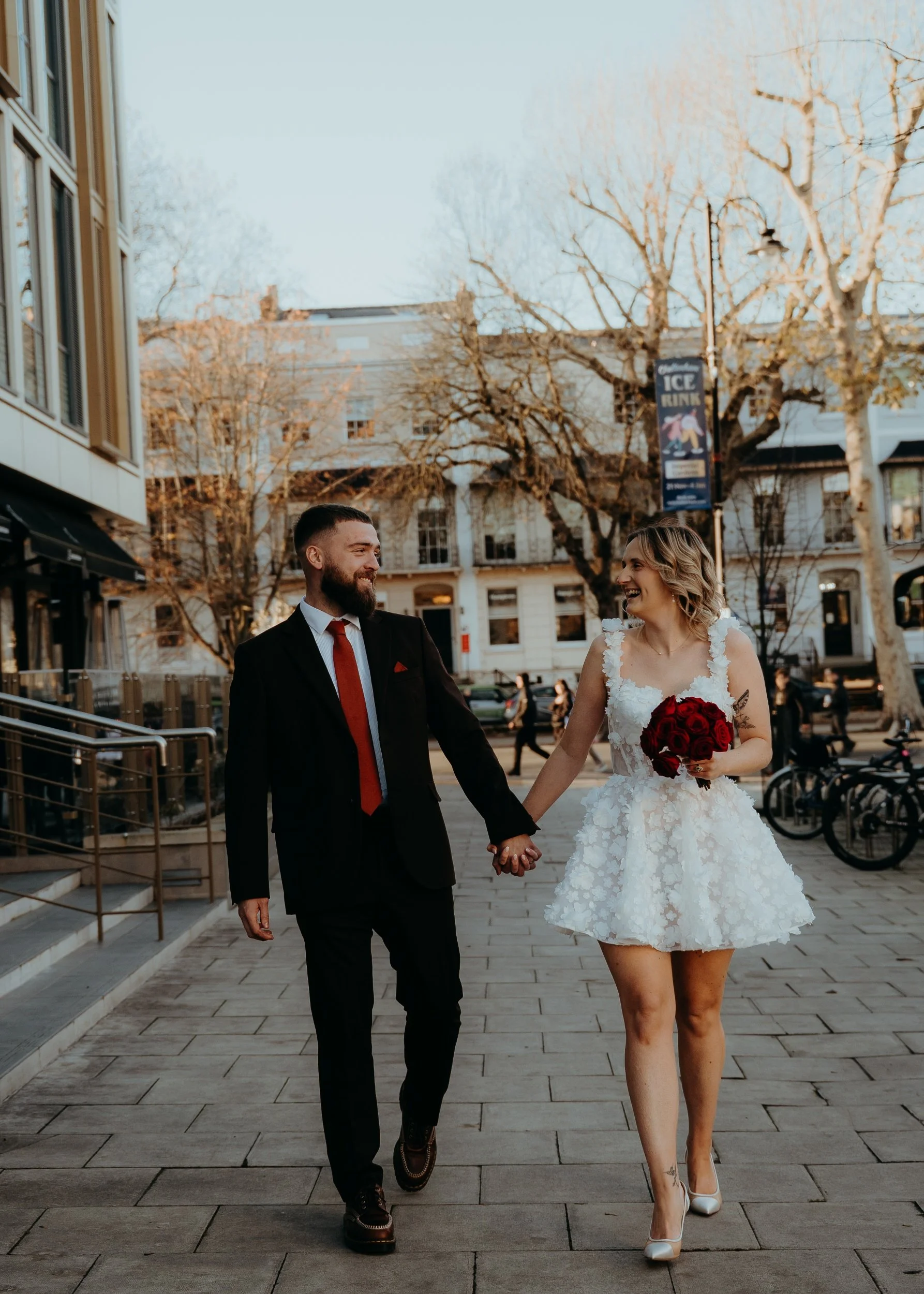 A newlywed couple holding hands and smiling while walking on a city sidewalk during the daytime. The woman is wearing a white wedding dress and holding a bouquet of red roses, and the man is in a black suit with a red tie.