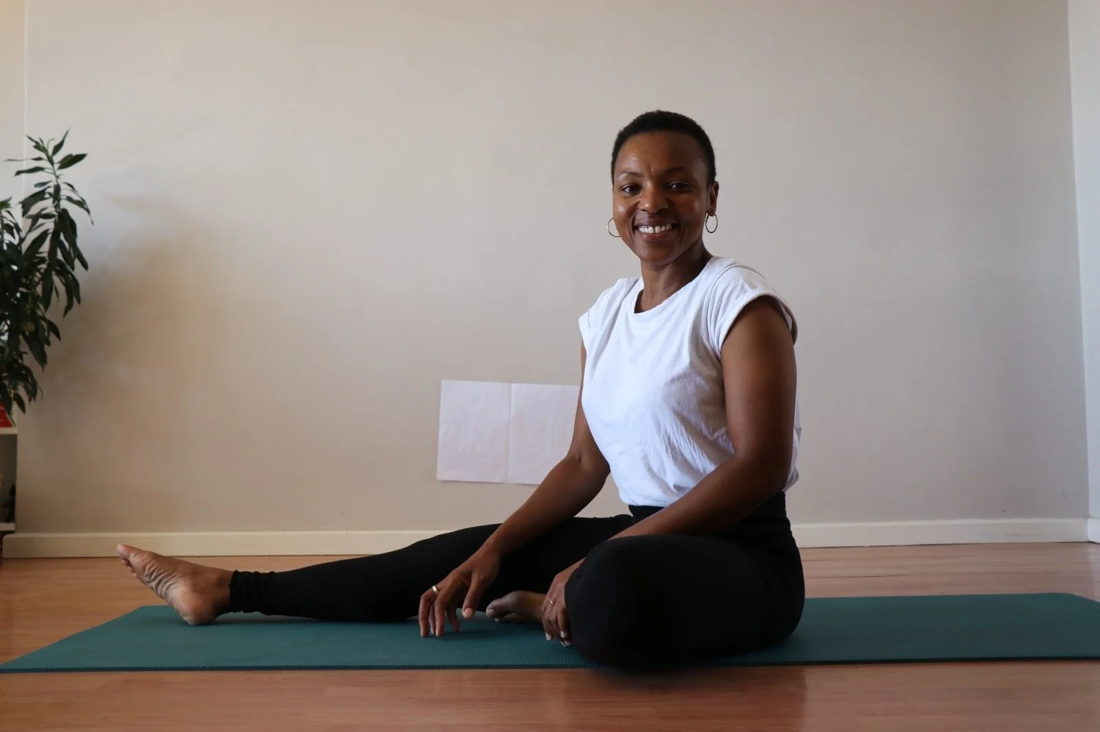 Woman sitting on yoga mat in a yoga pose, smiling, in a room with hardwood floor and a plant in the background.