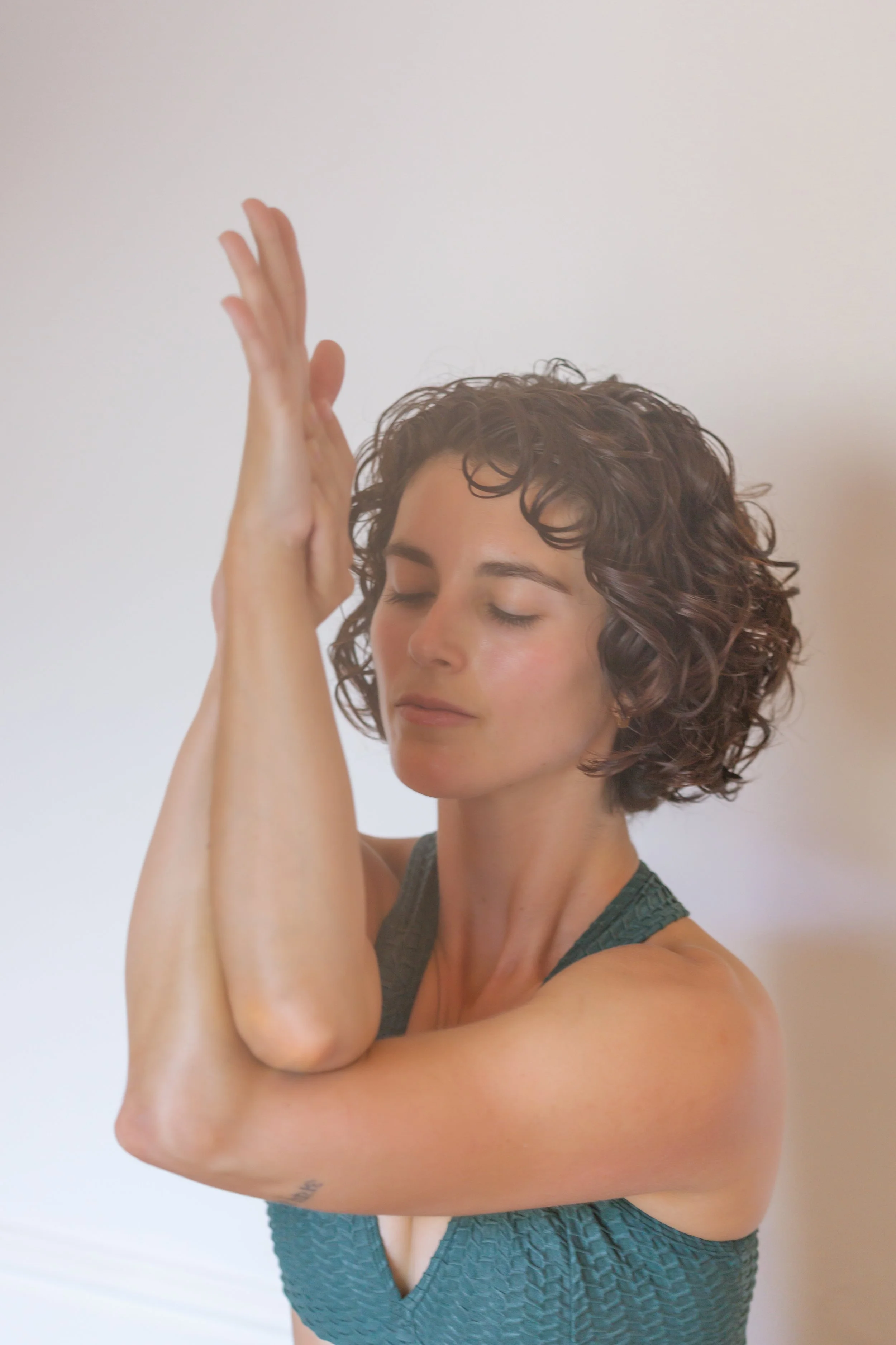 A woman with curly short brown hair practicing yoga in front of a plain white wall, with eyes closed and hands in prayer position above her head.