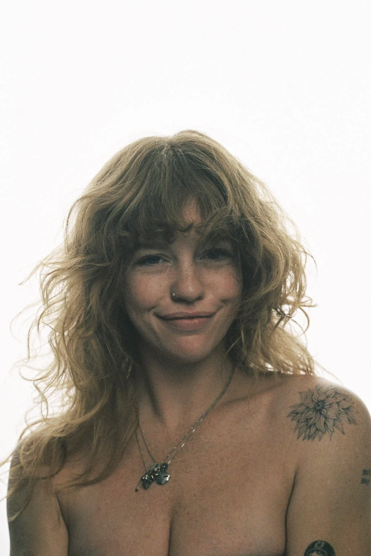 A woman with curly blond hair, a nose ring, a tattoo of a sunflower on her shoulder, and a floral tattoo on her arm, smiling and looking at the camera against a plain white background.