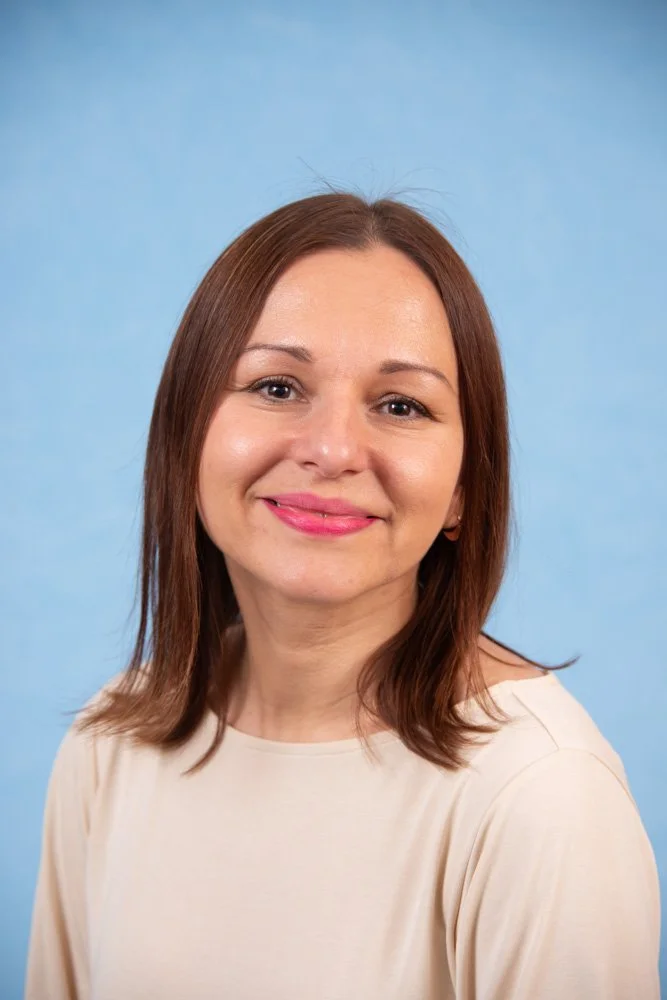 A woman with shoulder length brown hair wearing a cream top smiles in front of a blue background