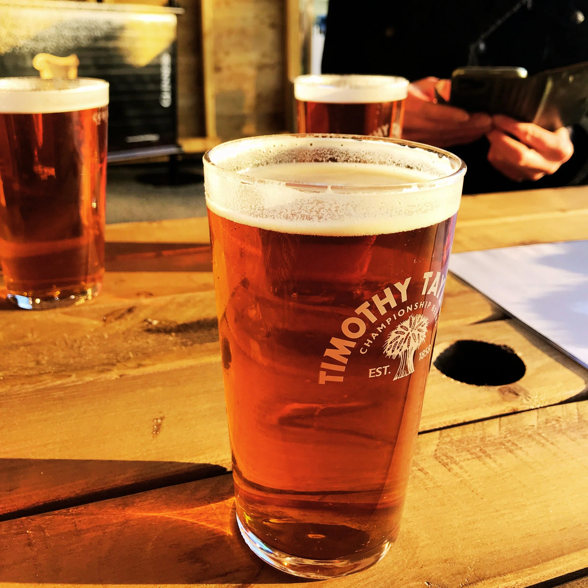 Three pints of beer on a wooden table with someone looking at their phone in the background