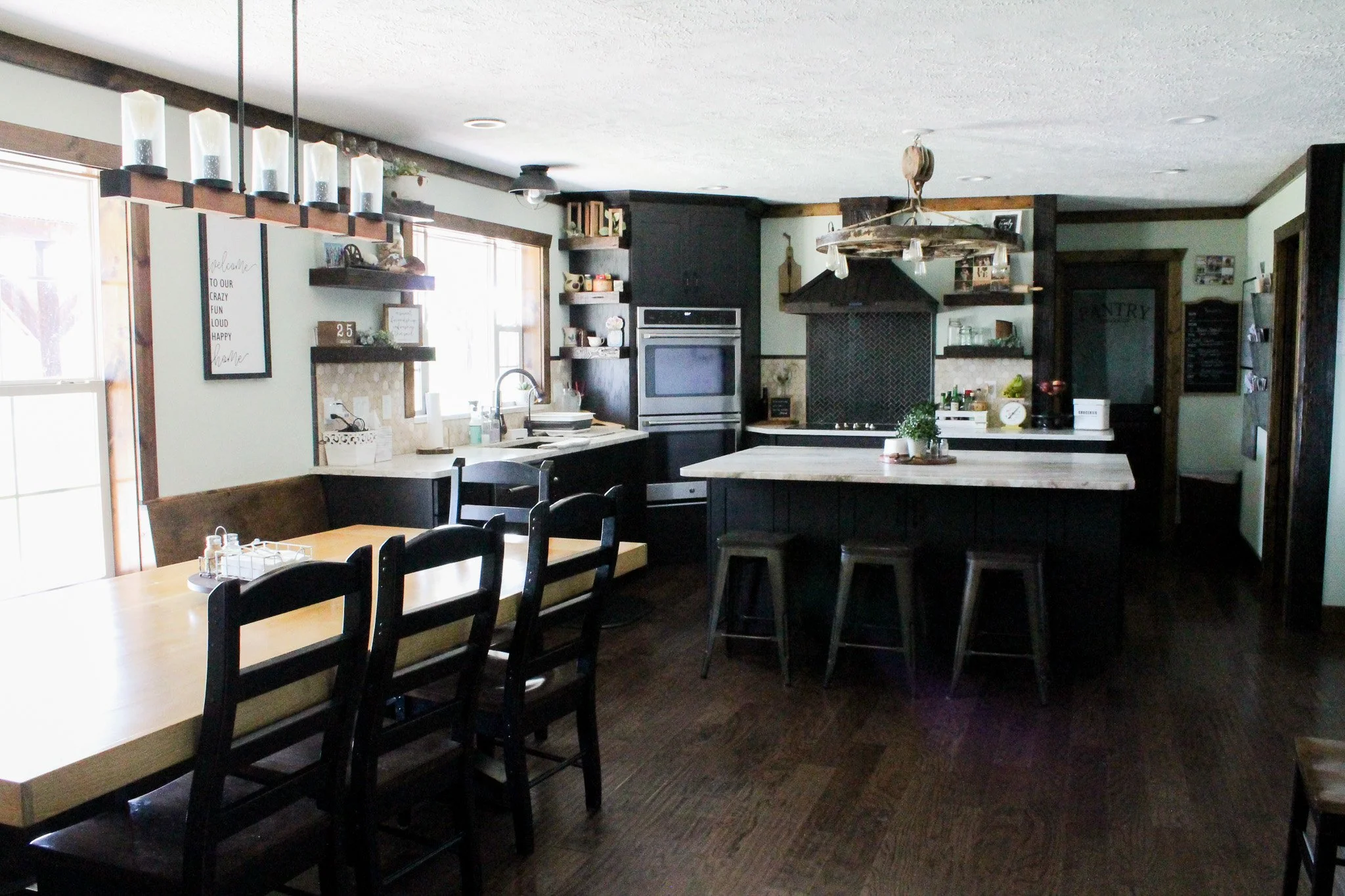 Right-angled Wide view of the kitchen showing it in its entirety. From right to left, kitchen island, stove top, double oven unit, kitchen sink, and window, rectangle kitchen table.