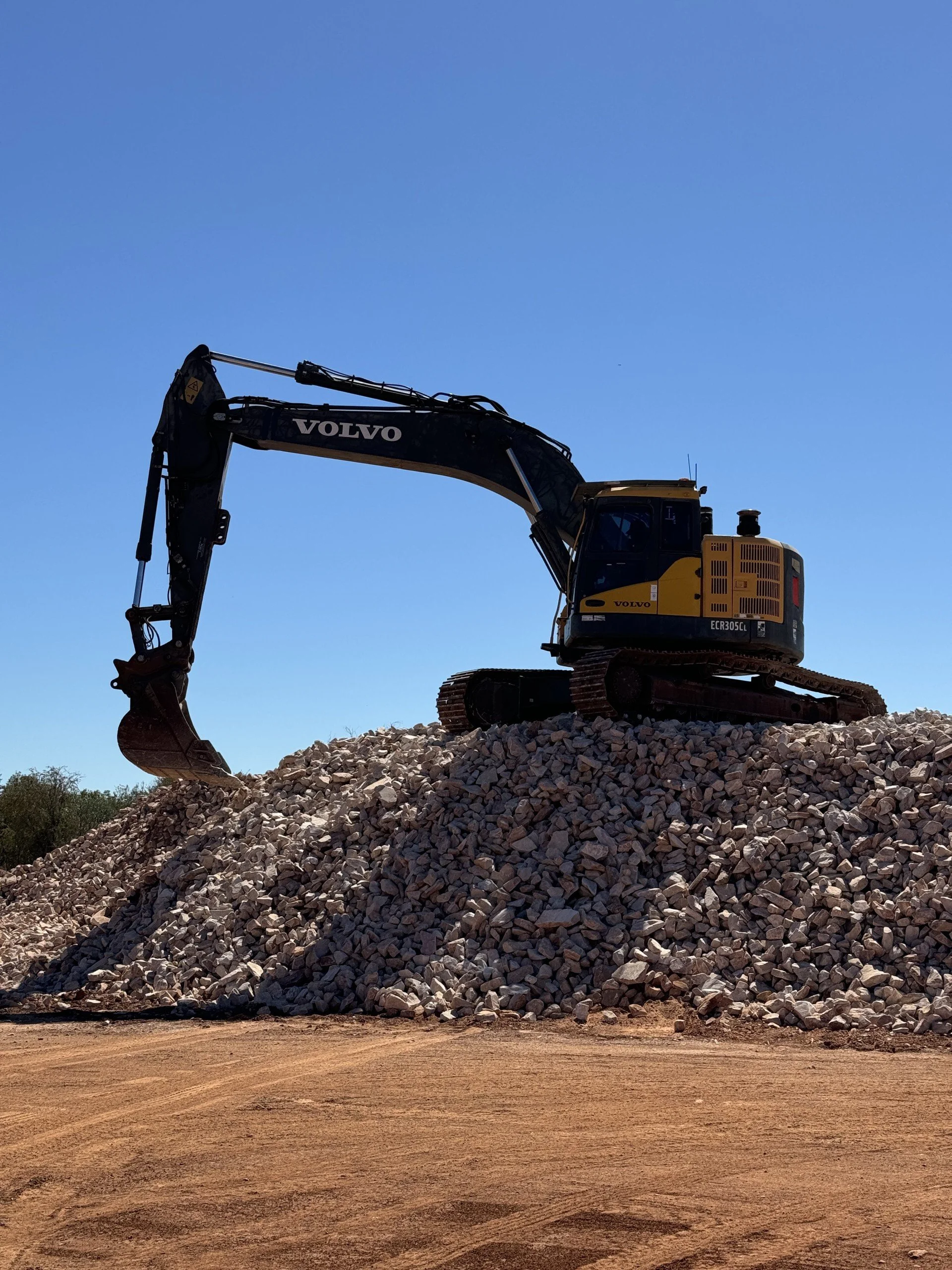 A Volvo excavator positioned on a large pile of rocks at a construction site under a clear blue sky.