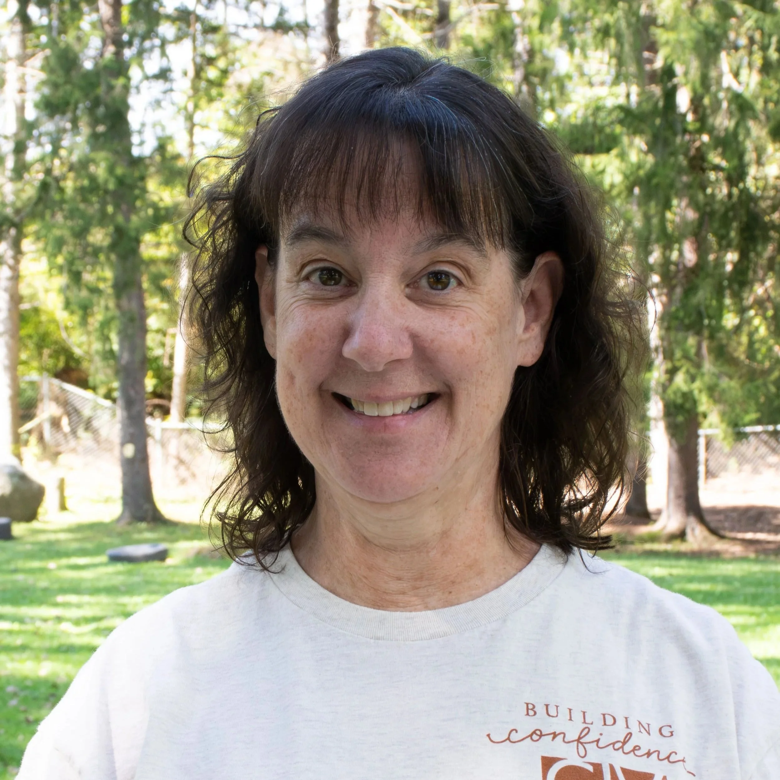 A woman with brown, curly hair and a bright smile, wearing a black blouse with red and beige floral patterns, standing against a grey background.
