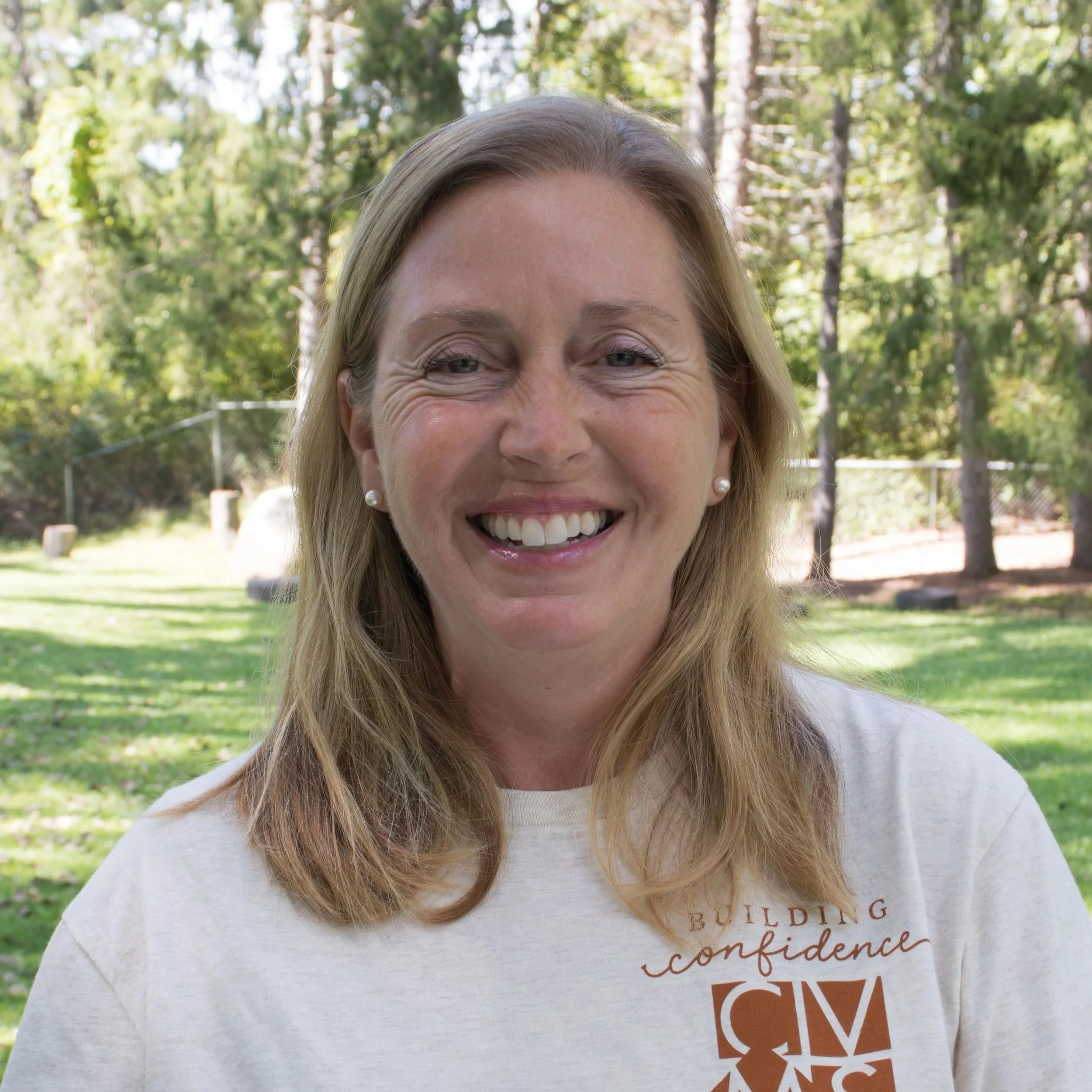 Close-up portrait of a smiling woman with blonde hair, wearing pearl earrings, and a brown top, against a gray background.