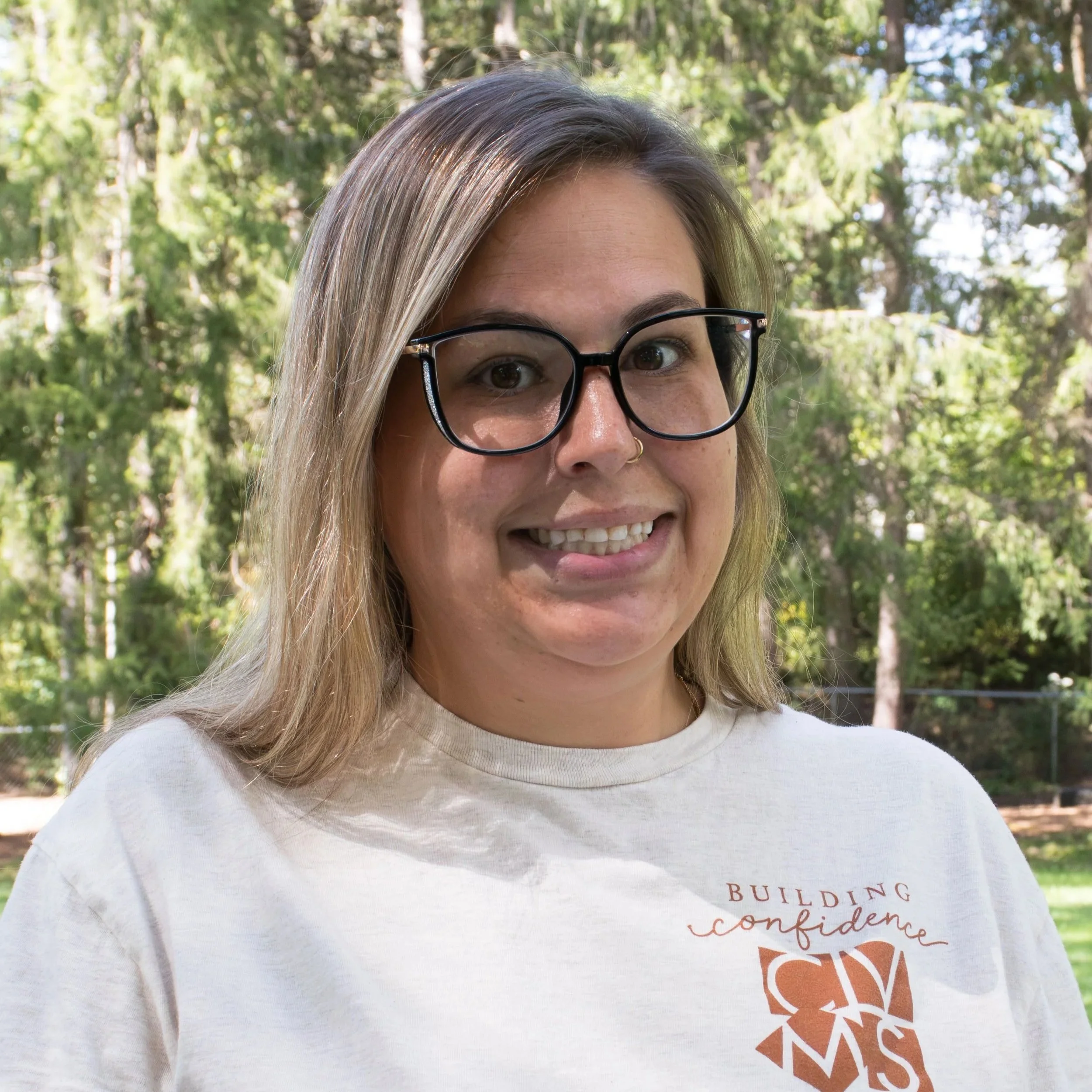 A young woman with long brown hair, light skin, and a nose piercing, smiling and wearing a brown sweater against a gray background.