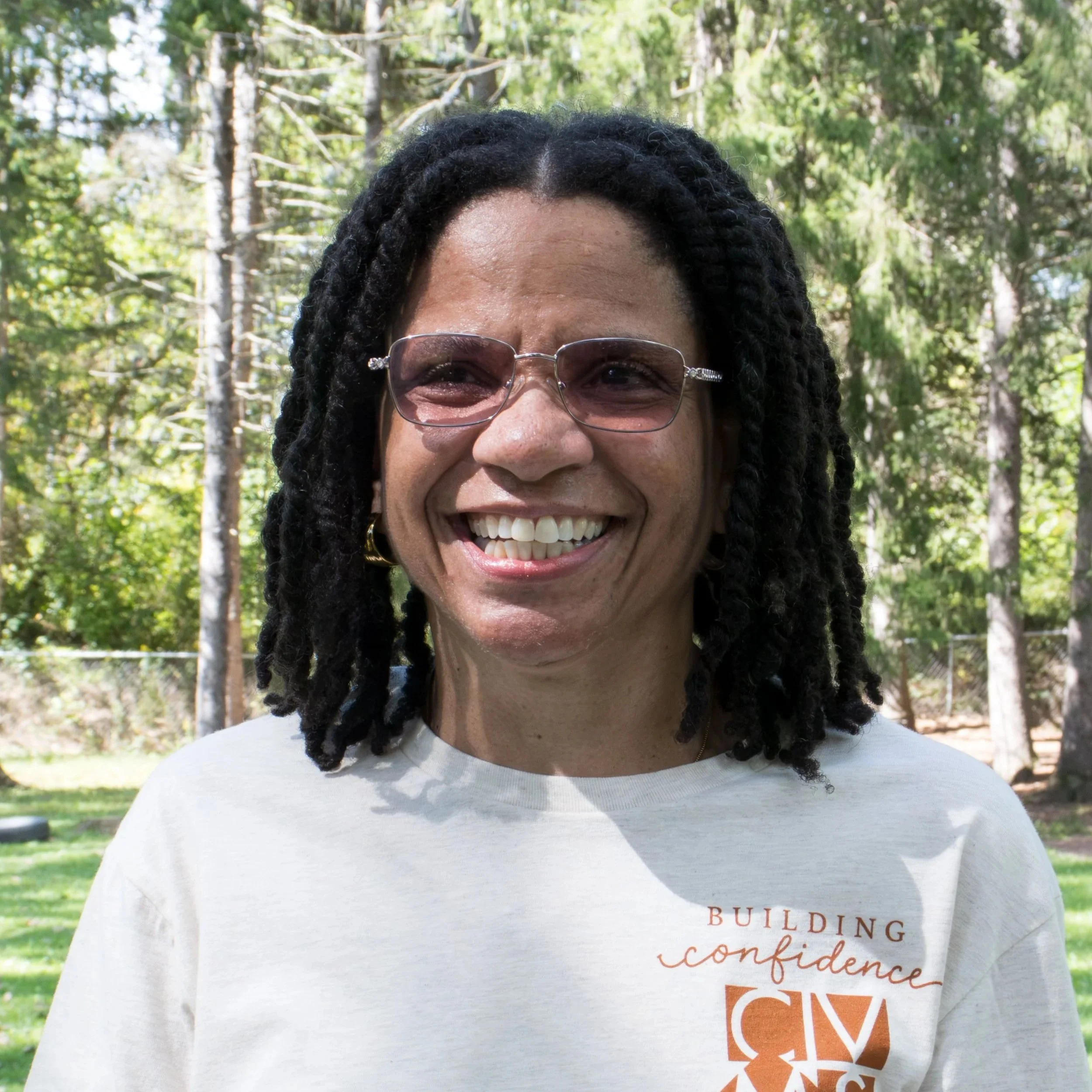 Portrait of a smiling woman with short curly hair, hoop earrings, and a colorful patterned blouse against a gray background.