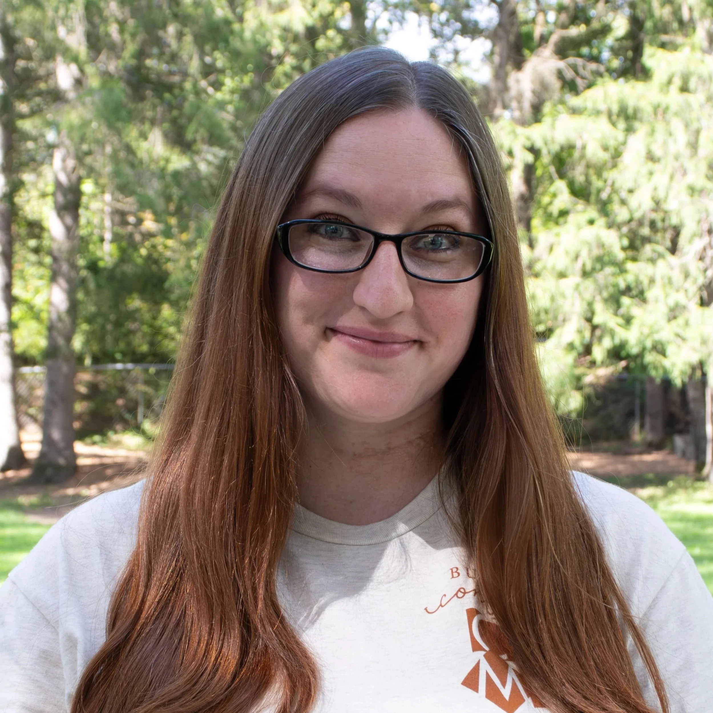 A woman with straight, dark red hair and black glasses, wearing a striped shirt, smiling against a gray background.