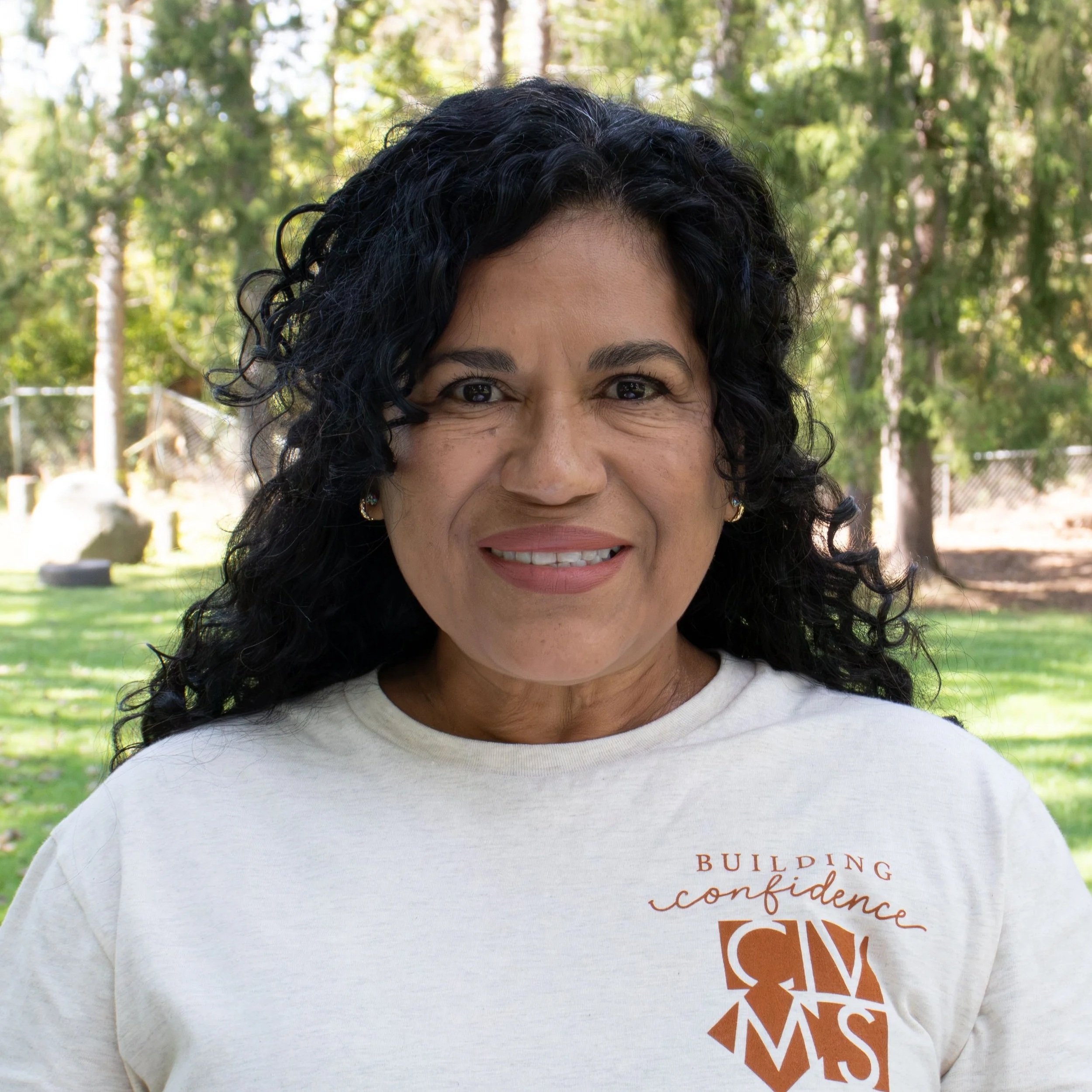A woman with dark, curly hair smiling at the camera, wearing earrings and a black top.