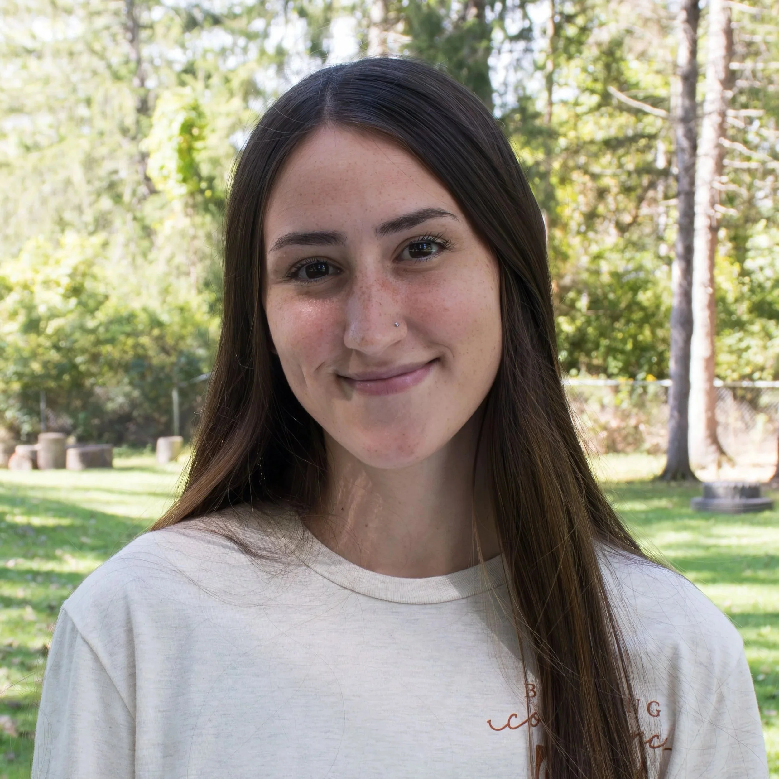Young woman with long brown hair, wearing a white turtleneck and rust-colored sweater, smiling against a gray background.