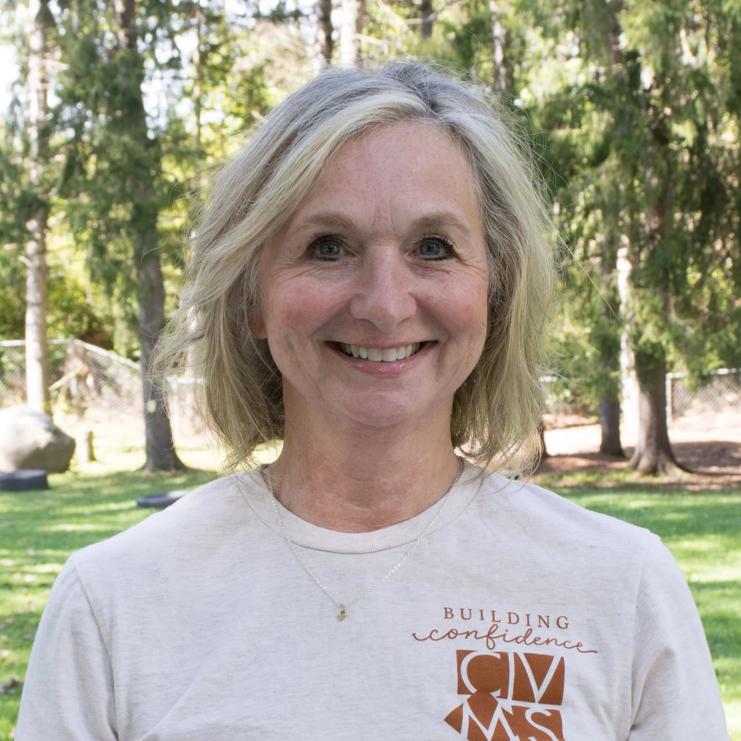 A woman with shoulder-length blonde hair, smiling, wearing a patterned blouse and beige cardigan, standing against a neutral gray background.