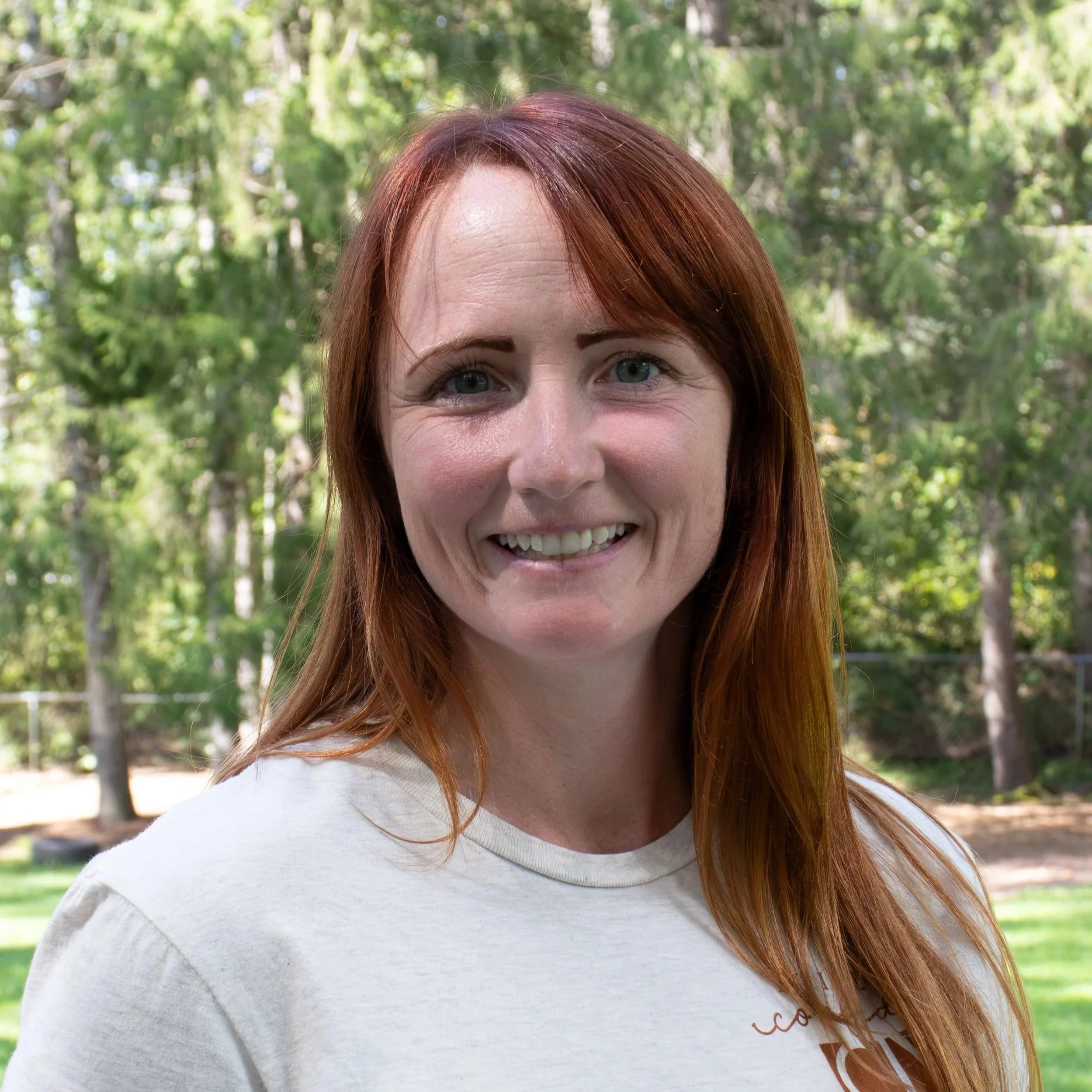 A woman with long red hair, smiling and wearing earrings, dressed in a black top, with a gray background.