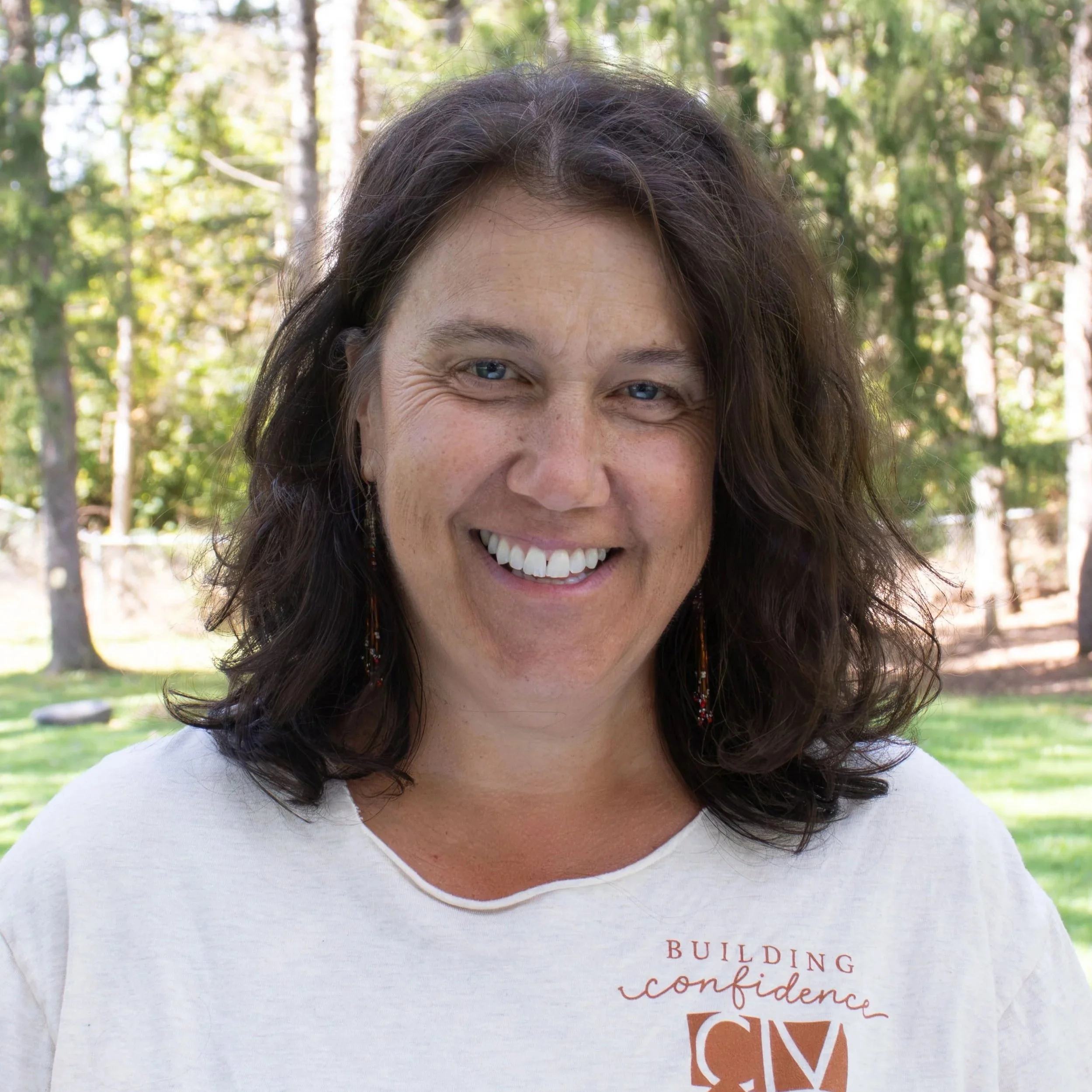 Portrait of a woman with shoulder-length brown hair, smiling, wearing a dark collared shirt and dangling earrings, in front of a neutral gray background.