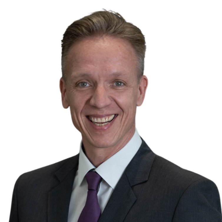 Smiling man in a dark suit, white shirt, and purple tie against a white background.