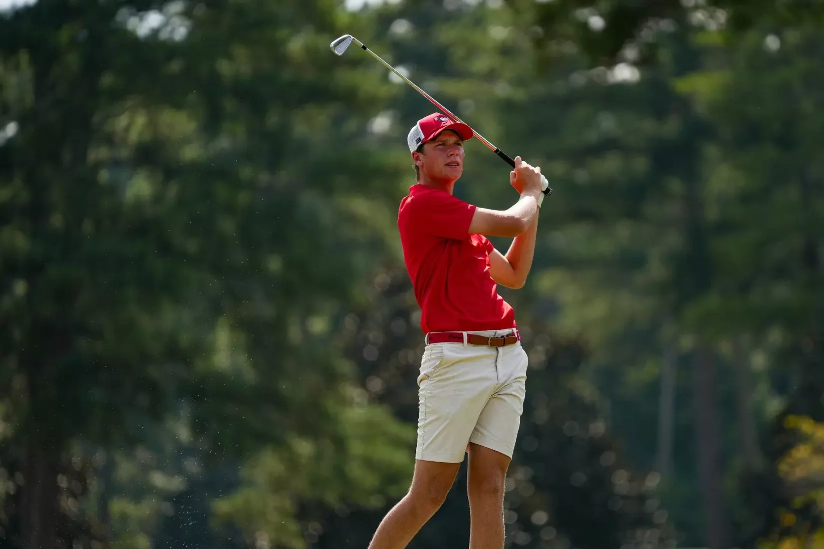 A young male golfer in a red shirt, white shorts, and a red cap swinging a golf club on a golf course with trees in the background.