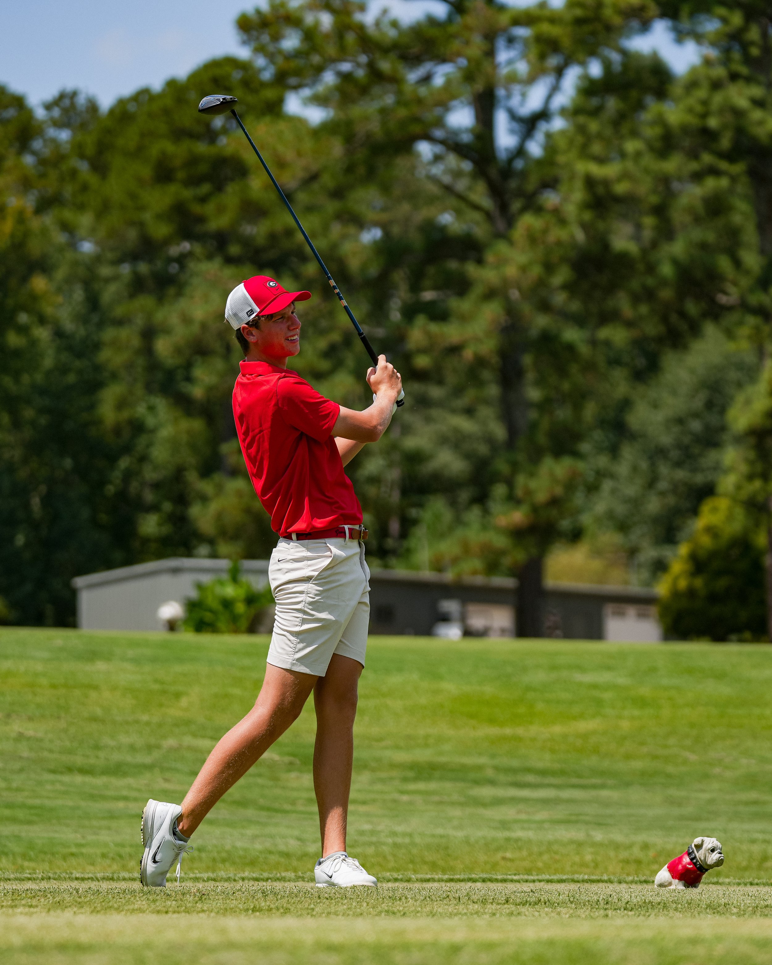 A young man in a red shirt, khaki shorts, and a baseball cap, playing golf on a sunny day. He is standing on a golf course with trees in the background, holding a golf club. A dog wearing a red coat is sitting on the grass nearby.