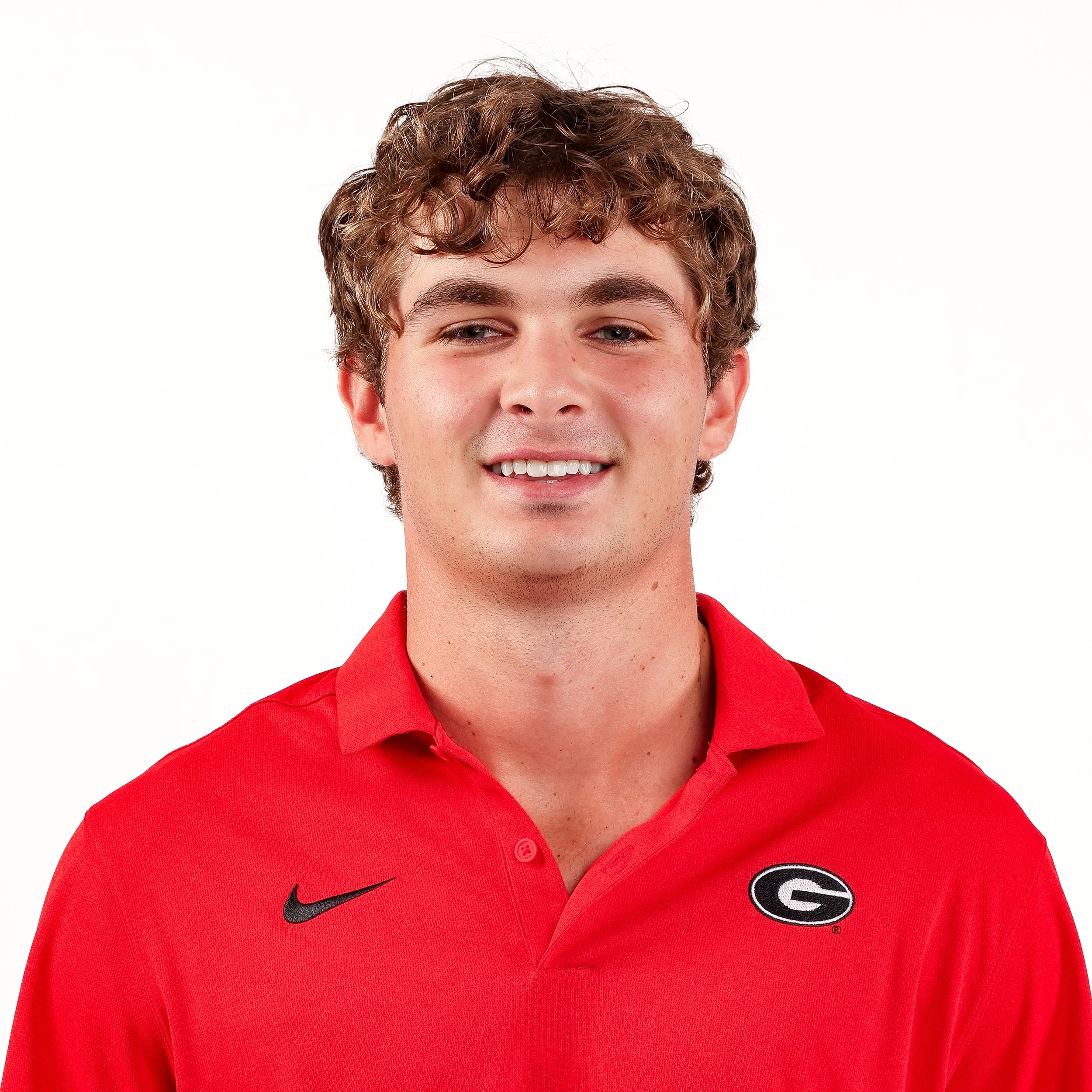 A young man with curly brown hair and fair skin wearing a red collared shirt with the University of Georgia logo and Nike swoosh, standing against a plain white background.