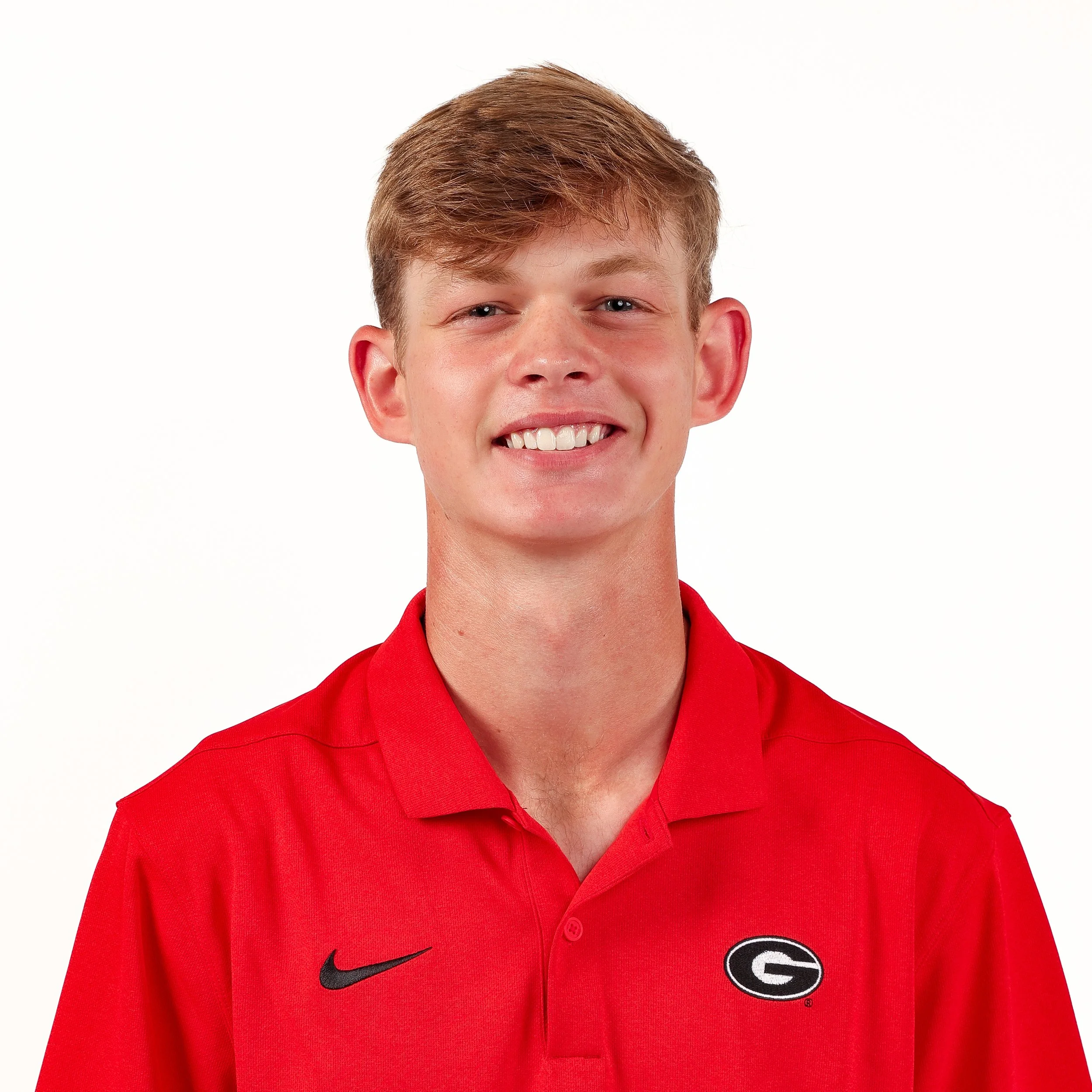 Close-up of a young man with short red hair, wearing a red collared shirt with a black Nike logo and a Georgia Bulldogs logo on it, smiling against a white background.