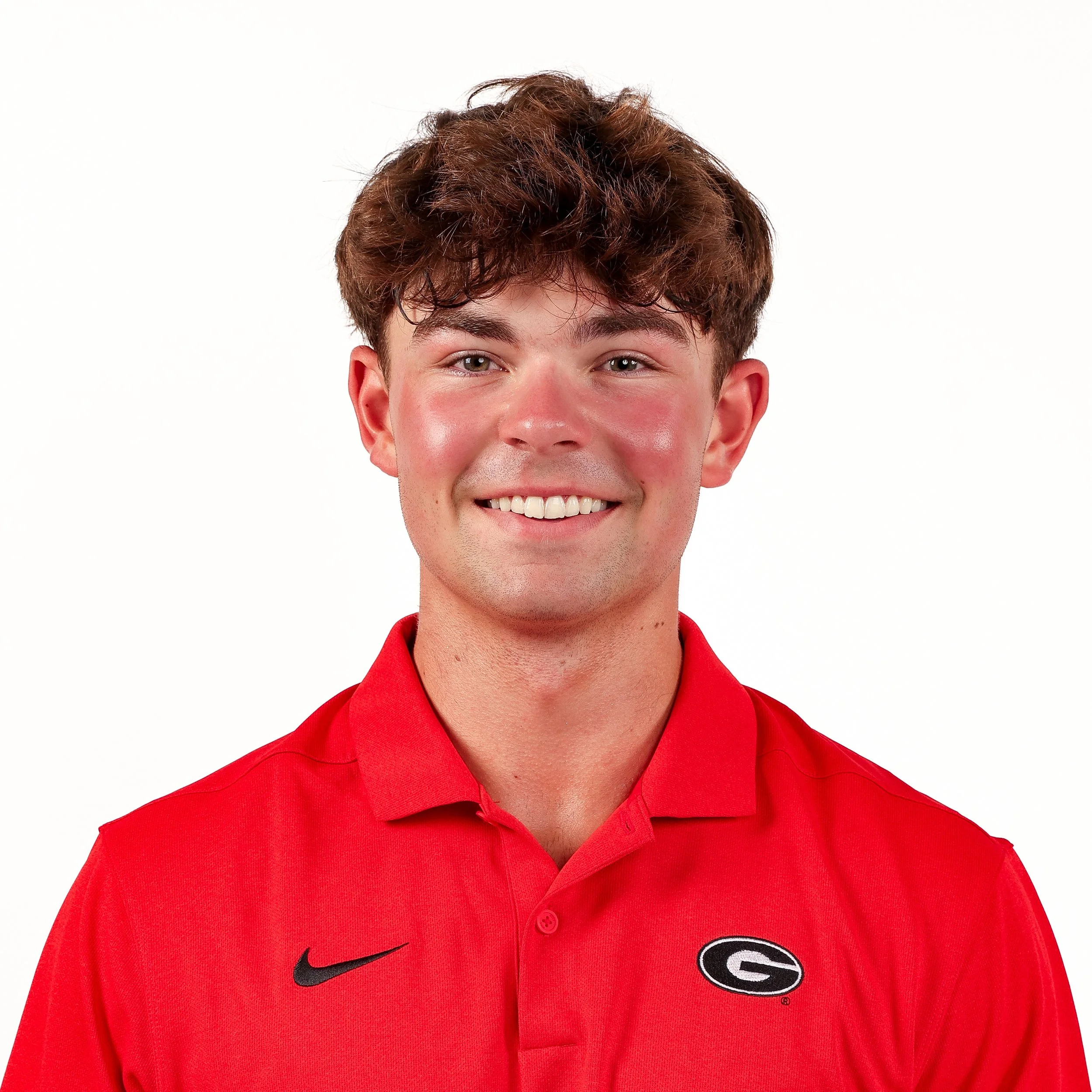 Close-up of a young man's face with curly brown hair and a big smile, wearing a red sports polo shirt with Gator logo and Nike swoosh on a plain white background.