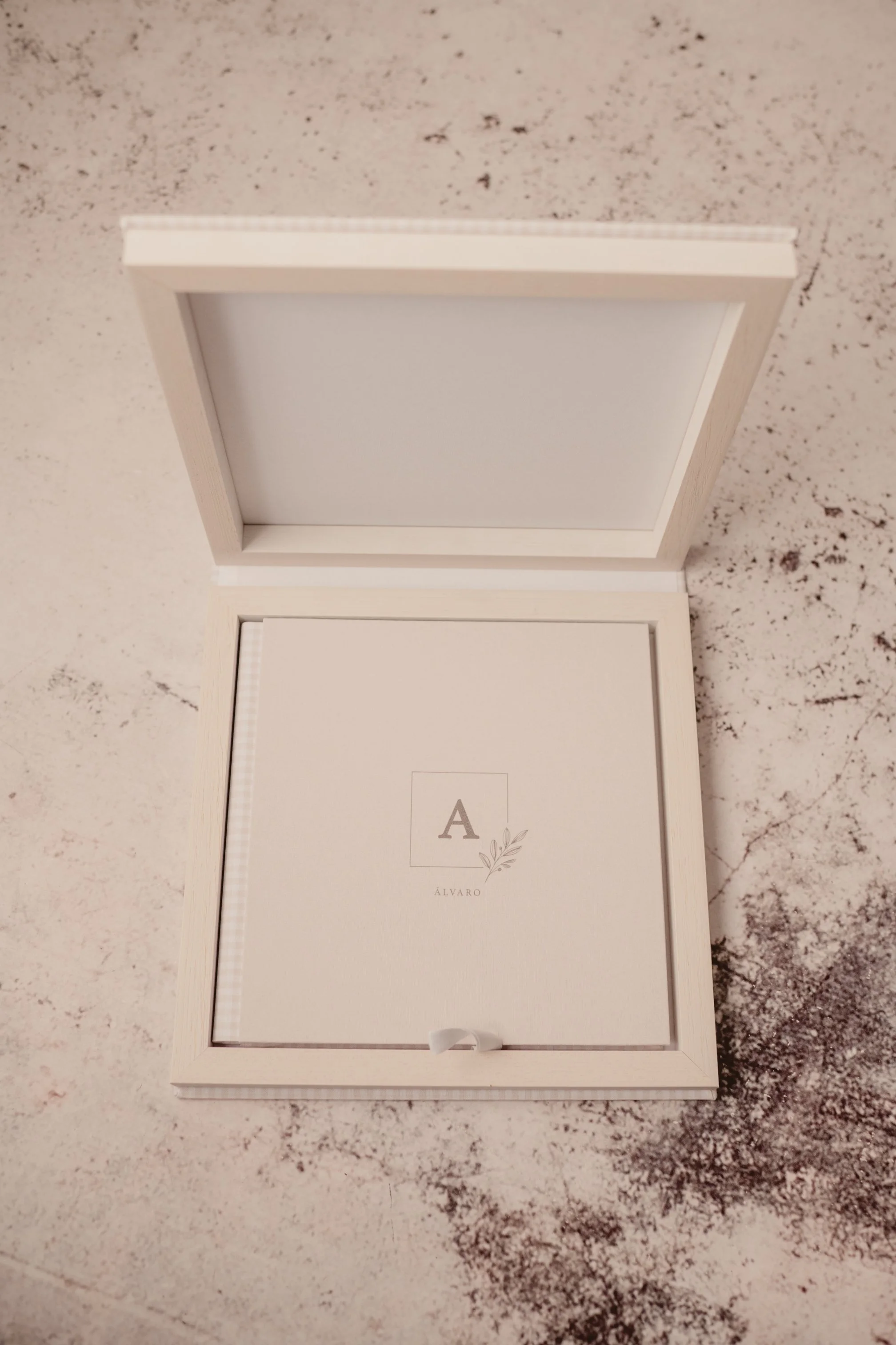 Caja de presentación blanca con la letra 'A' y el nombre 'Álvaro' en el centro, decorada con una rama de olivo, sobre una superficie de piedra beige con manchas oscuras.