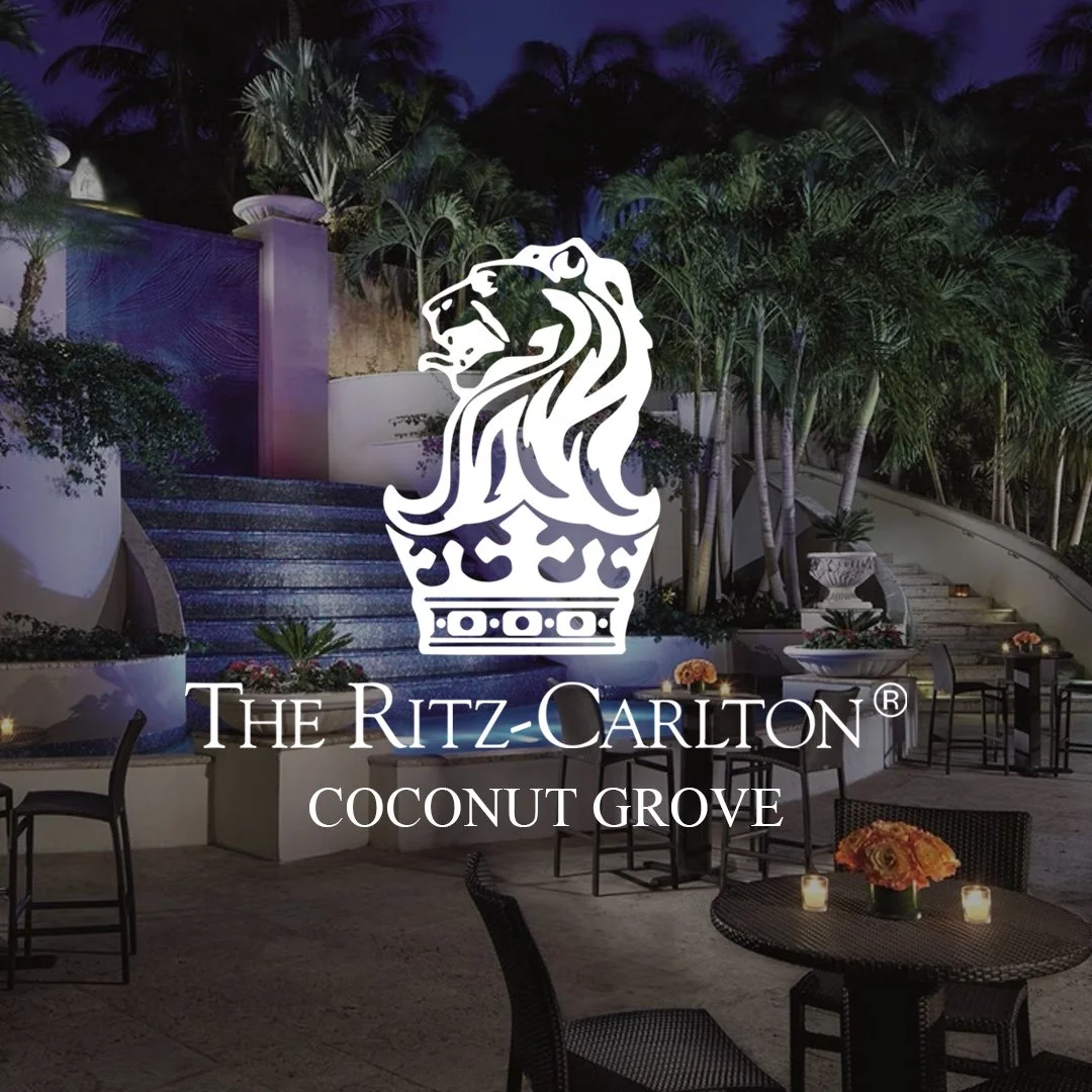 Outdoor patio of The Ritz-Carlton Coconut Grove with tables, chairs, and palm trees, featuring hotel logo.