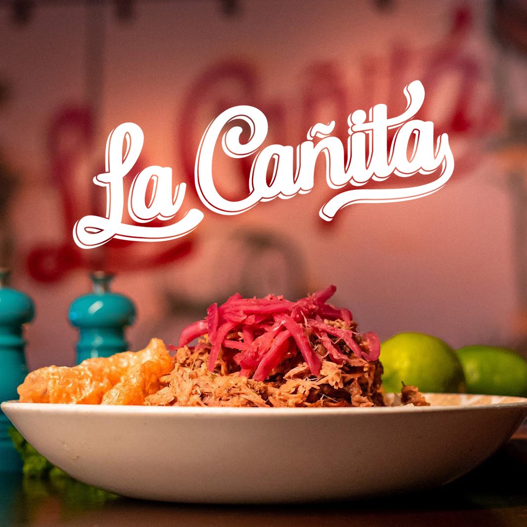 Plate of shredded meat topped with pickled onions, alongside fried pork rinds and limes, with blurred teal salt and pepper shakers in the background. The words 'La Cañita' appear prominently above the dish.