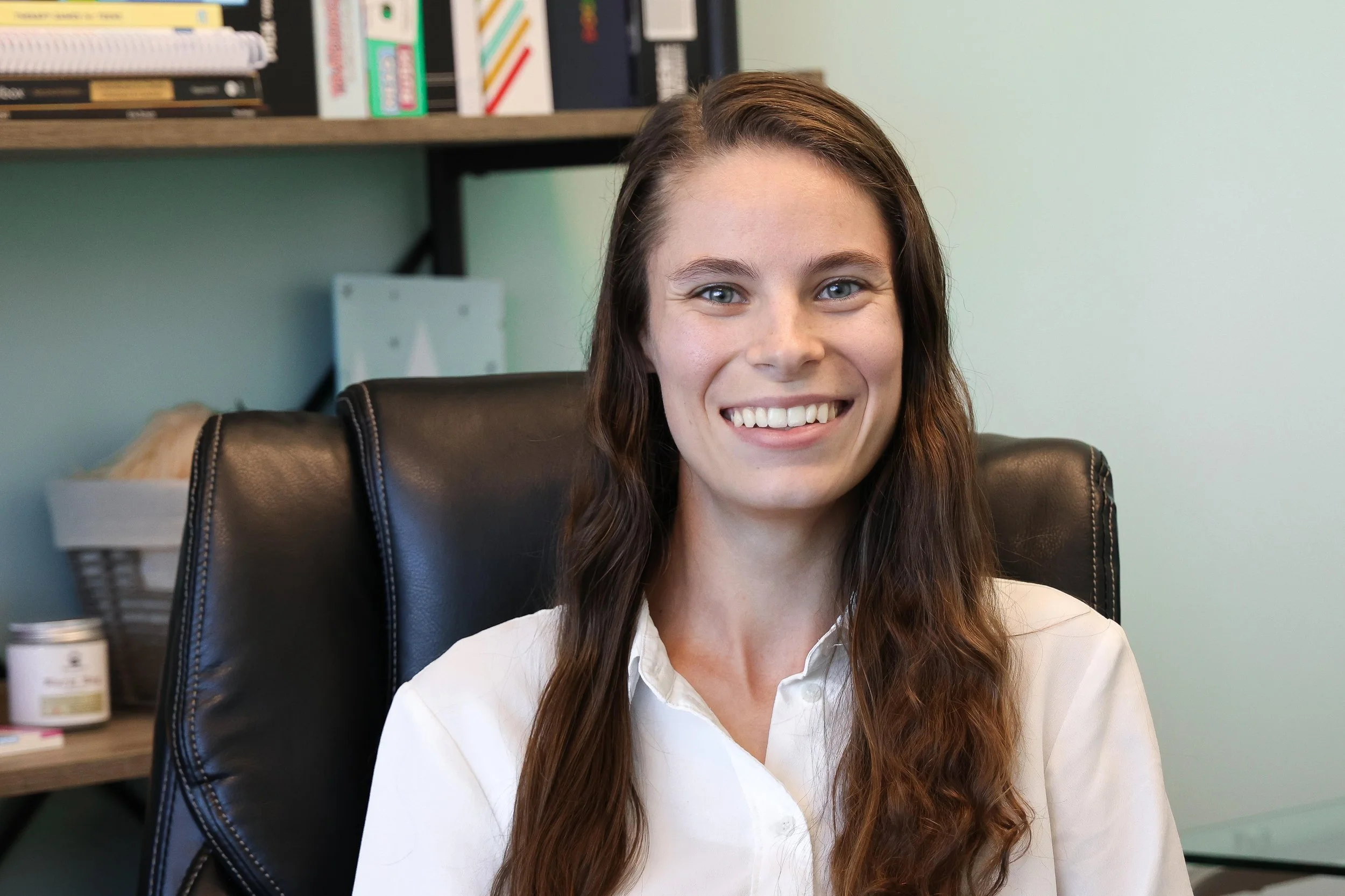 A young woman with long brown hair and blue eyes sitting in a black leather office chair, smiling at the camera, in an office setting with a light-colored wall and a wooden bookshelf with various books and supplies in the background.