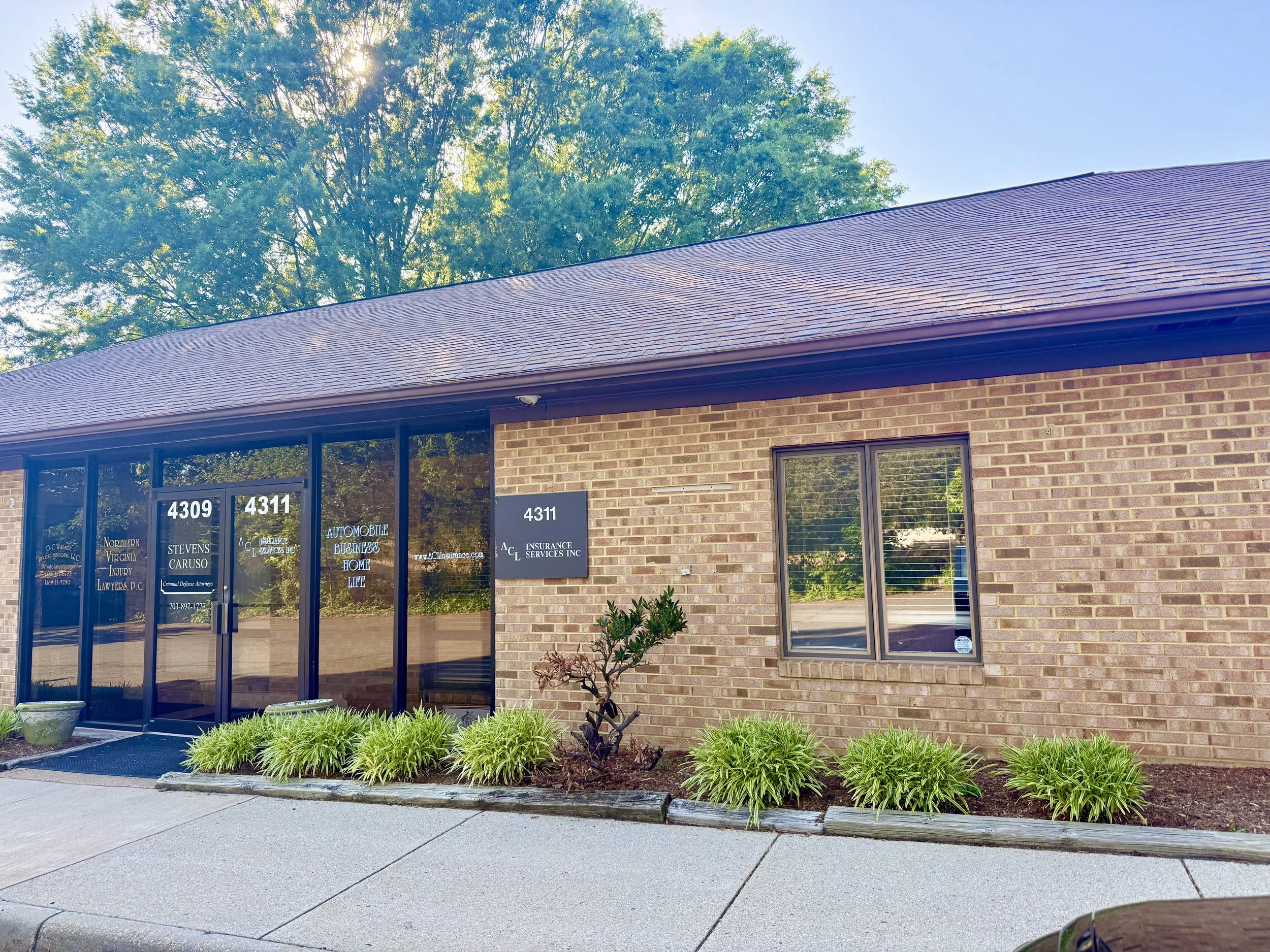 A brick commercial building with glass doors and windows, with three signs displaying business names and numbers, surrounded by green plants and grass, and a sidewalk in front.