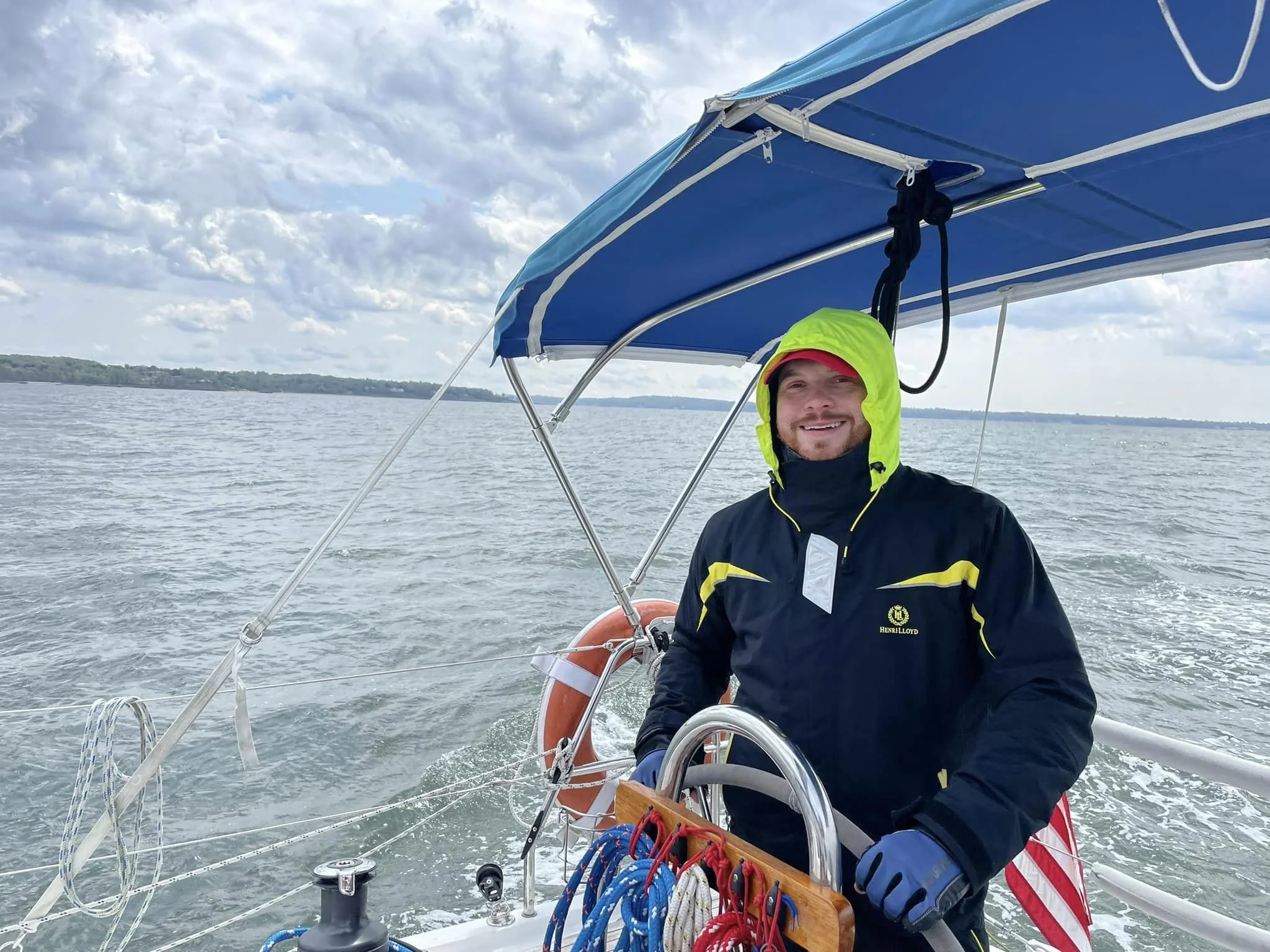 A man on a boat smiling, wearing a yellow and black waterproof jacket and a hood, with a death star emblem on the jacket, on a body of water with a cloudy sky in the background.