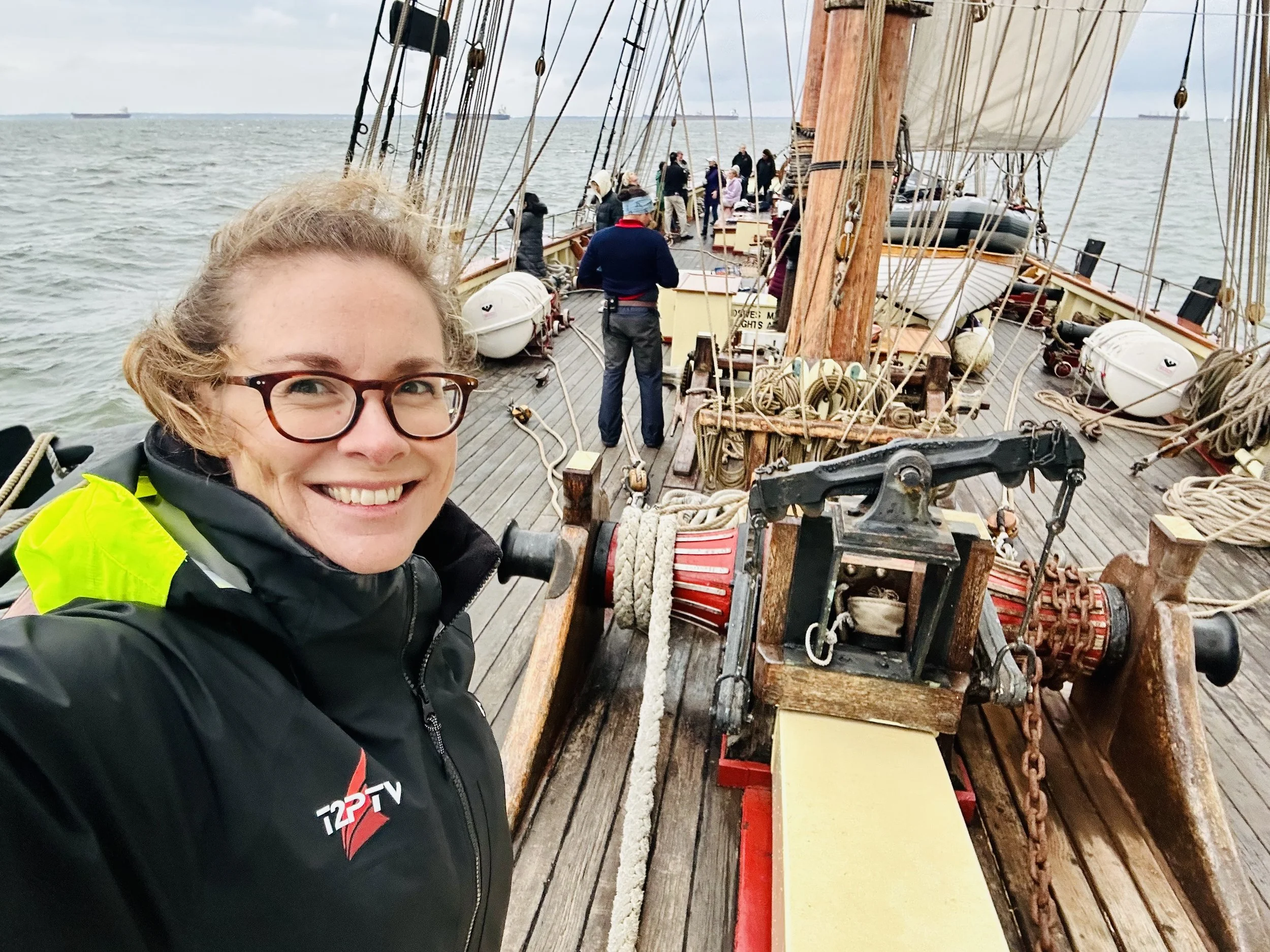 A woman with glasses taking a selfie on the deck of a sailing ship with rigging and crew members in the background on the ocean.