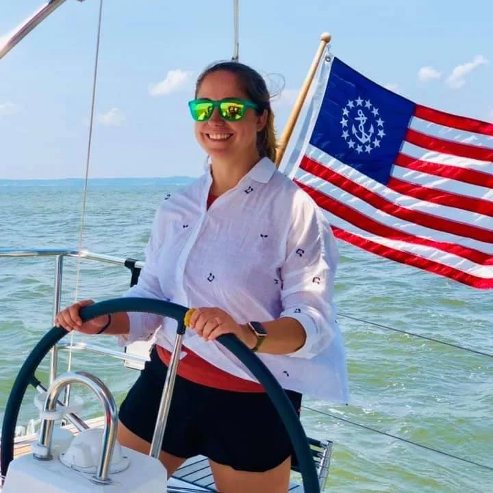 A woman smiling at the camera while steering a sailboat on the water, wearing sunglasses and a white shirt, with an American flag in the background.