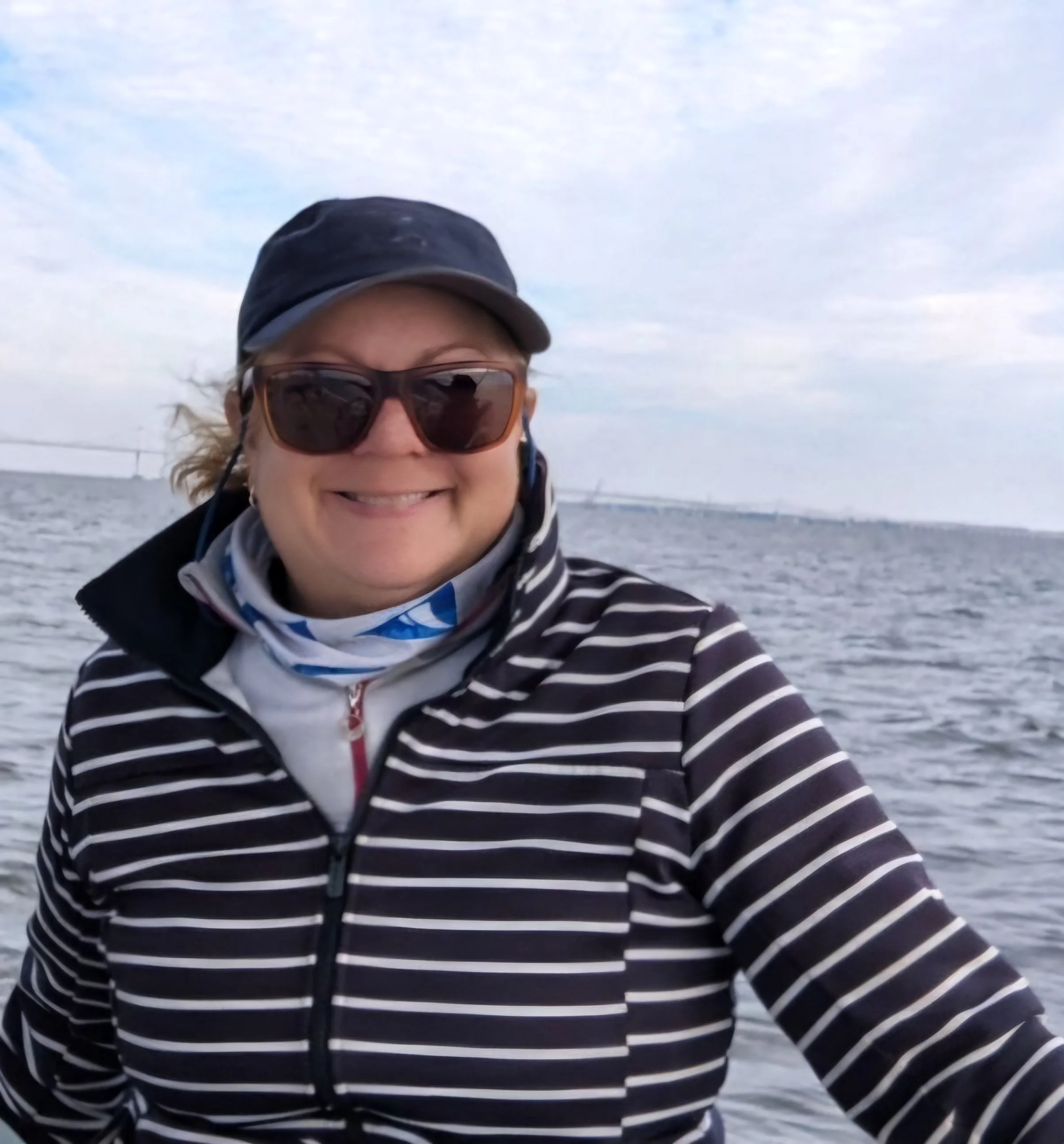 A woman smiling outdoors on a boat or near water, wearing sunglasses, a navy cap, a striped jacket, and a neck gaiter, with water and a cloudy sky in the background.