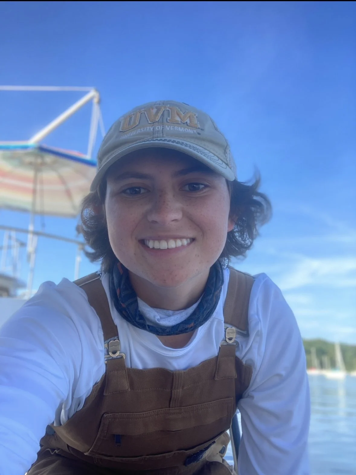 A young person smiling outdoors on a sunny day, wearing a UVM baseball cap, a white shirt, a brown life jacket, and a neck gaiter, with water and sailboats in the background.