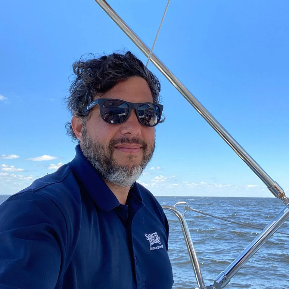 A man with dark hair and a beard wearing sunglasses and a navy blue collared shirt, sailing on open water under a bright blue sky with some clouds.