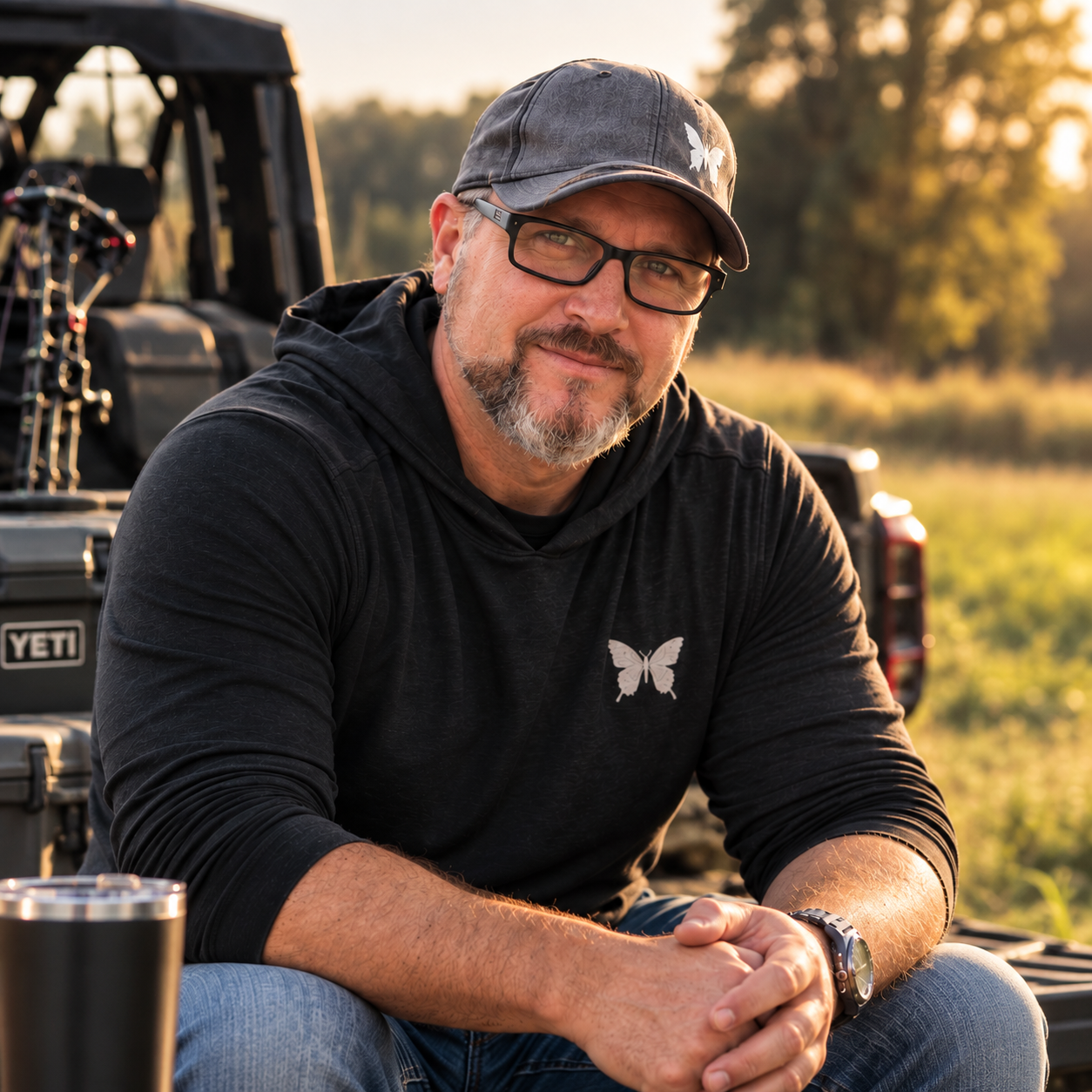 A middle-aged man with glasses and a beard sitting outdoors near a truck at sunset, wearing a black hoodie with a butterfly logo, and smiling at the camera.
