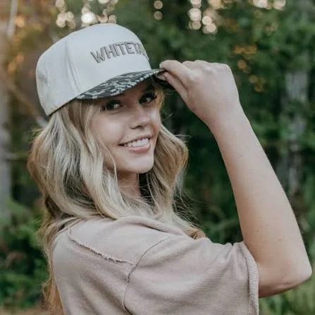 Young woman smiling outdoors, wearing a white baseball cap with 'WHITE' embroidered on it, beige t-shirt, in front of green trees.