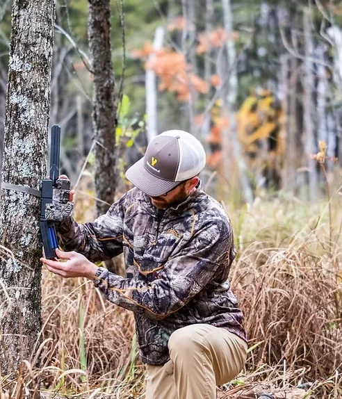 Man in camouflage jacket and baseball cap crouching in a forest, tending to a trail camera attached to a tree.