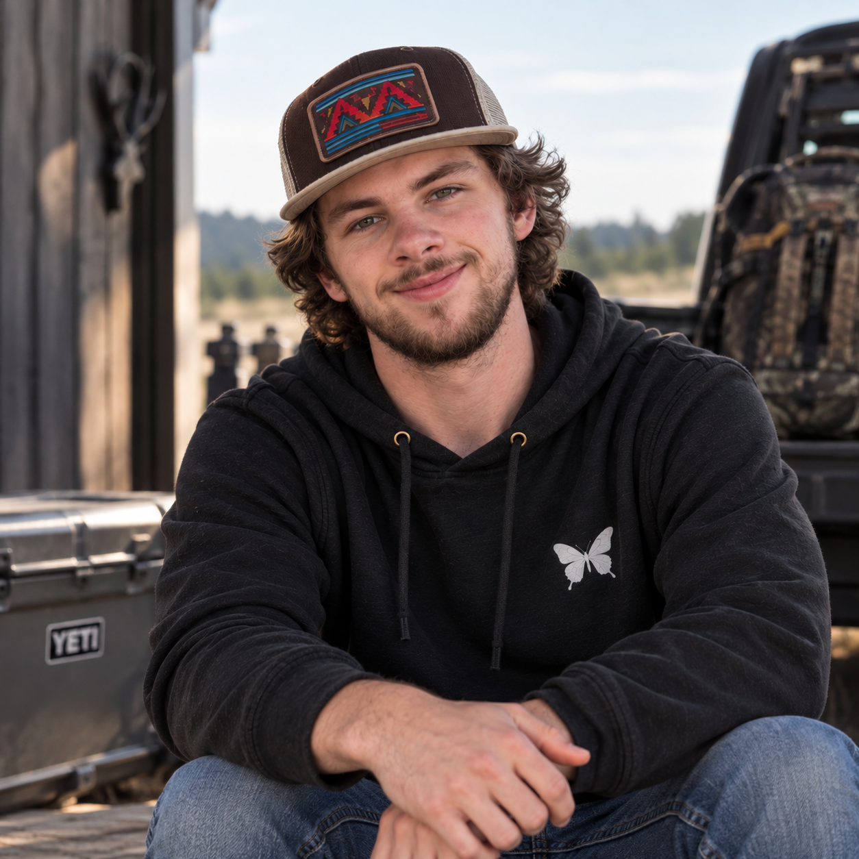 A young man with curly hair and a beard sitting outdoors, wearing a black hoodie with a butterfly logo and a brown cap with a mountain design. He is smiling and looking at the camera.