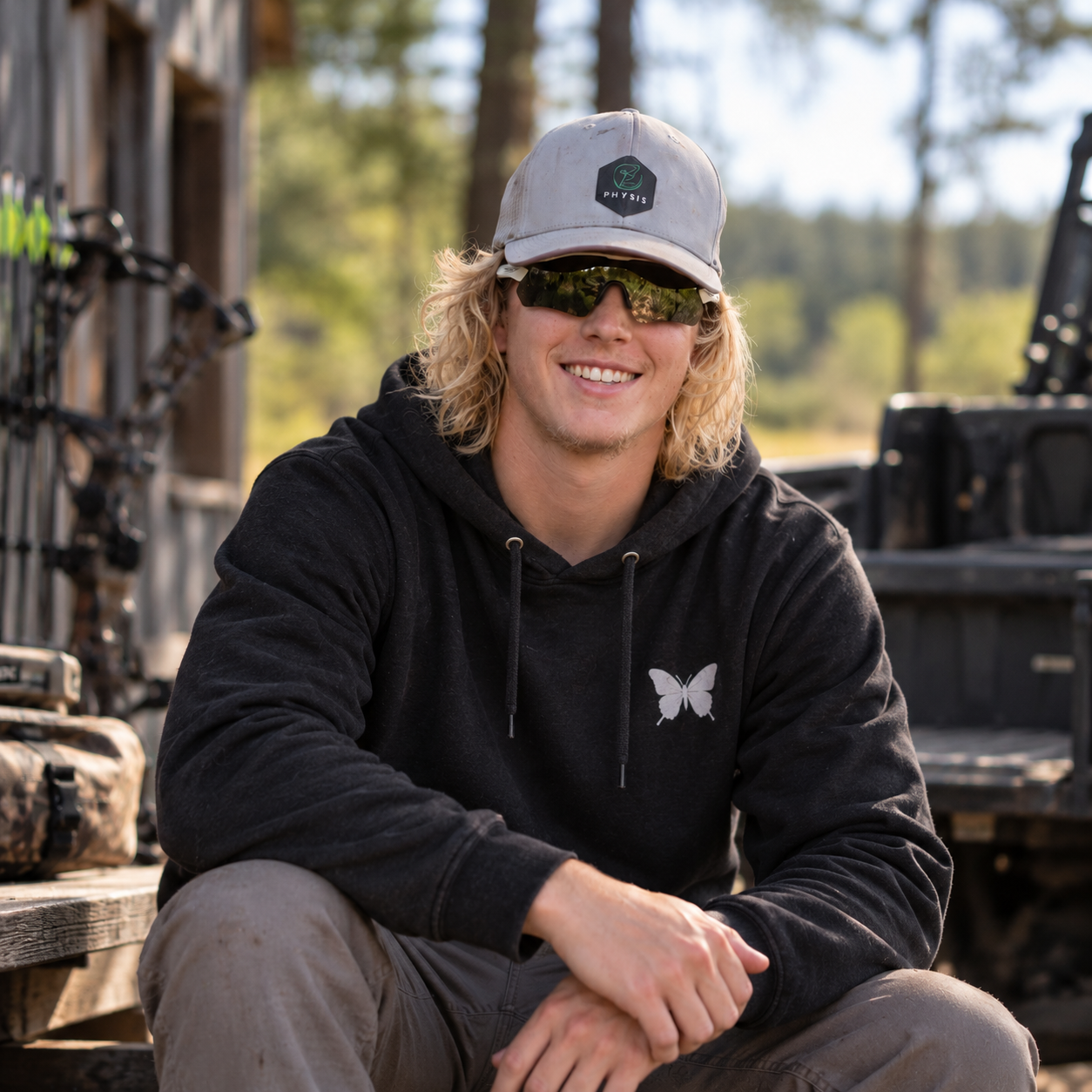 A young man with blonde curly hair wearing a gray cap with a 'PHYSIS' logo, dark sunglasses, a black hoodie with a butterfly logo, and beige pants, sitting outdoors with trees and a truck in the background, smiling.