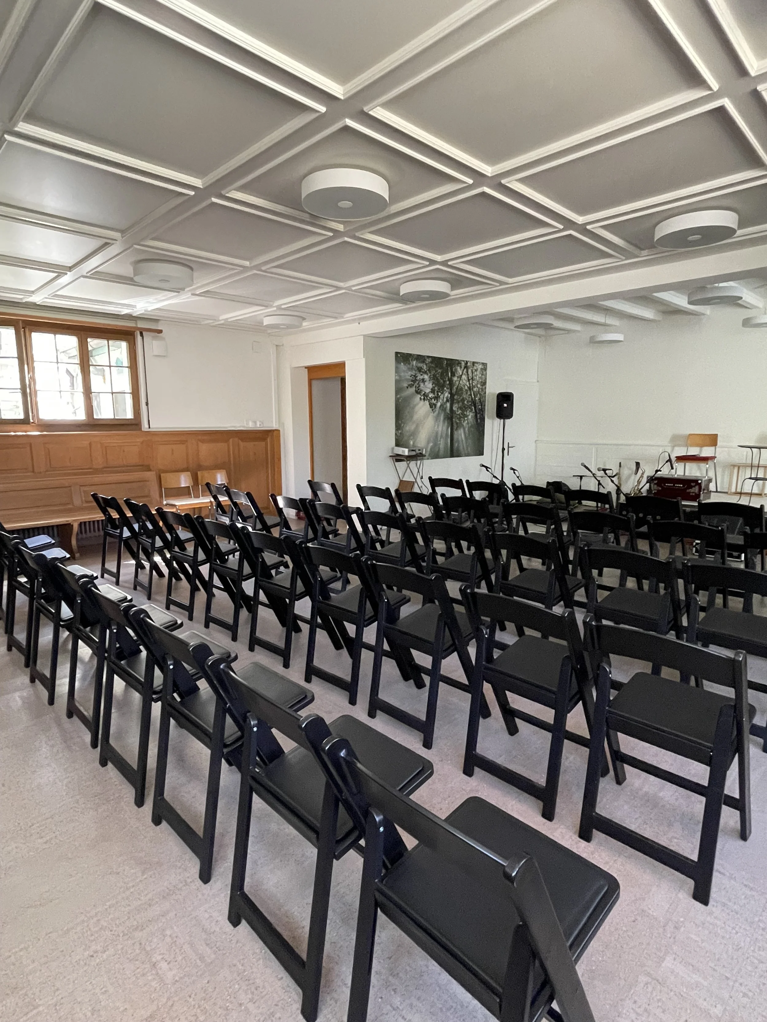 Ein Veranstaltungsraum mit schwarzen Stühlen in Reihen angeordnet. Im Hintergrund befindet sich ein Bild an der Wand und einige Musikinstrumente. Die Decke ist mit dekorativen Platten verziert.