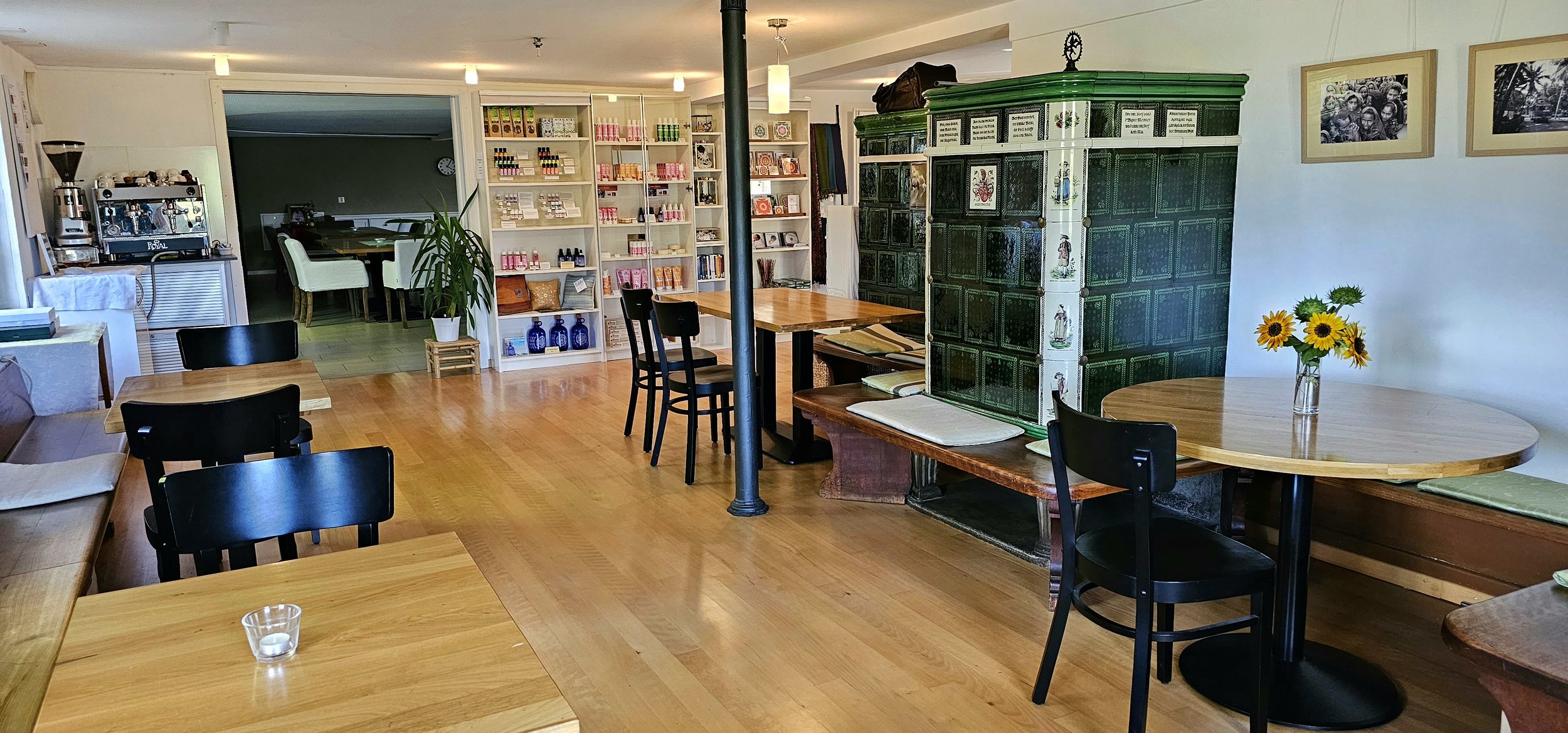 Gemütliches Café mit Holzmöbeln, einem Kachelofen, runden Tischen, Stühlen und einem Regal mit Produkten. Blumen in einer Vase auf einem Tisch.