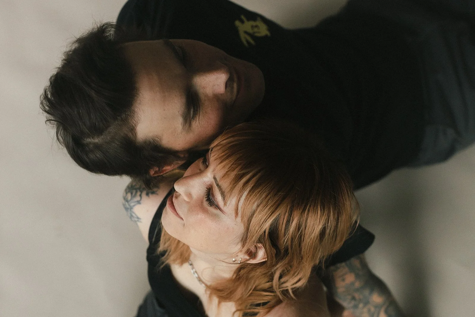 A young man and woman lying close together on the floor, with their faces touching and eyes closed in an intimate moment.