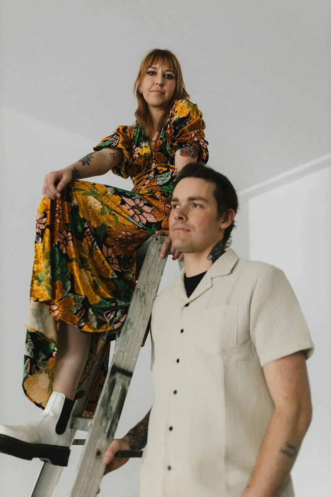 A woman with red hair and tattoos, wearing a colorful floral dress, is sitting on a ladder. A man with dark hair, wearing a cream-colored short-sleeve shirt, stands beside her, with a neutral expression against a plain white wall.