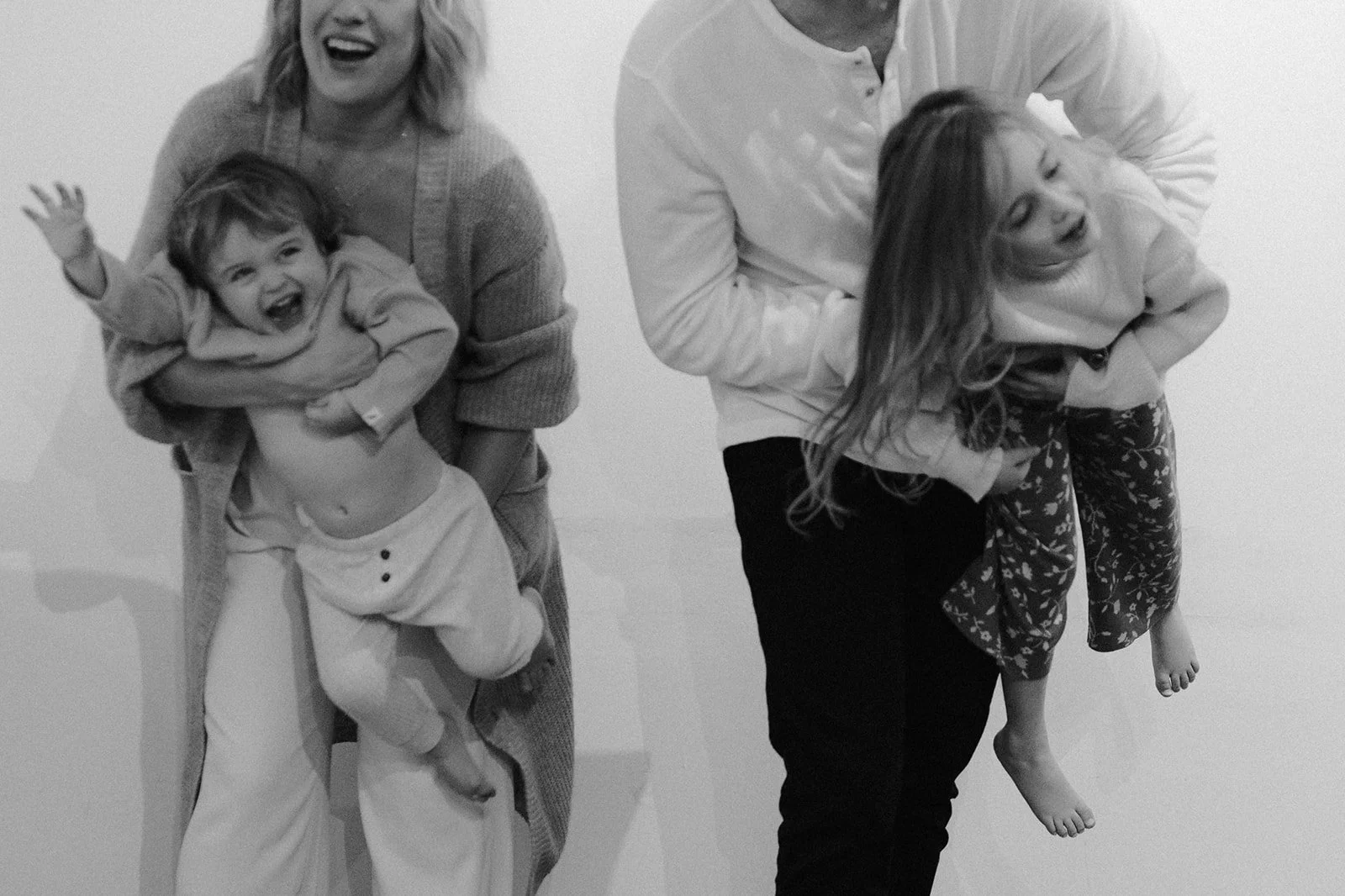 Two women each holding a laughing child, one child is a boy and the other a girl, in a playful piggyback pose, all smiling and appearing joyful.