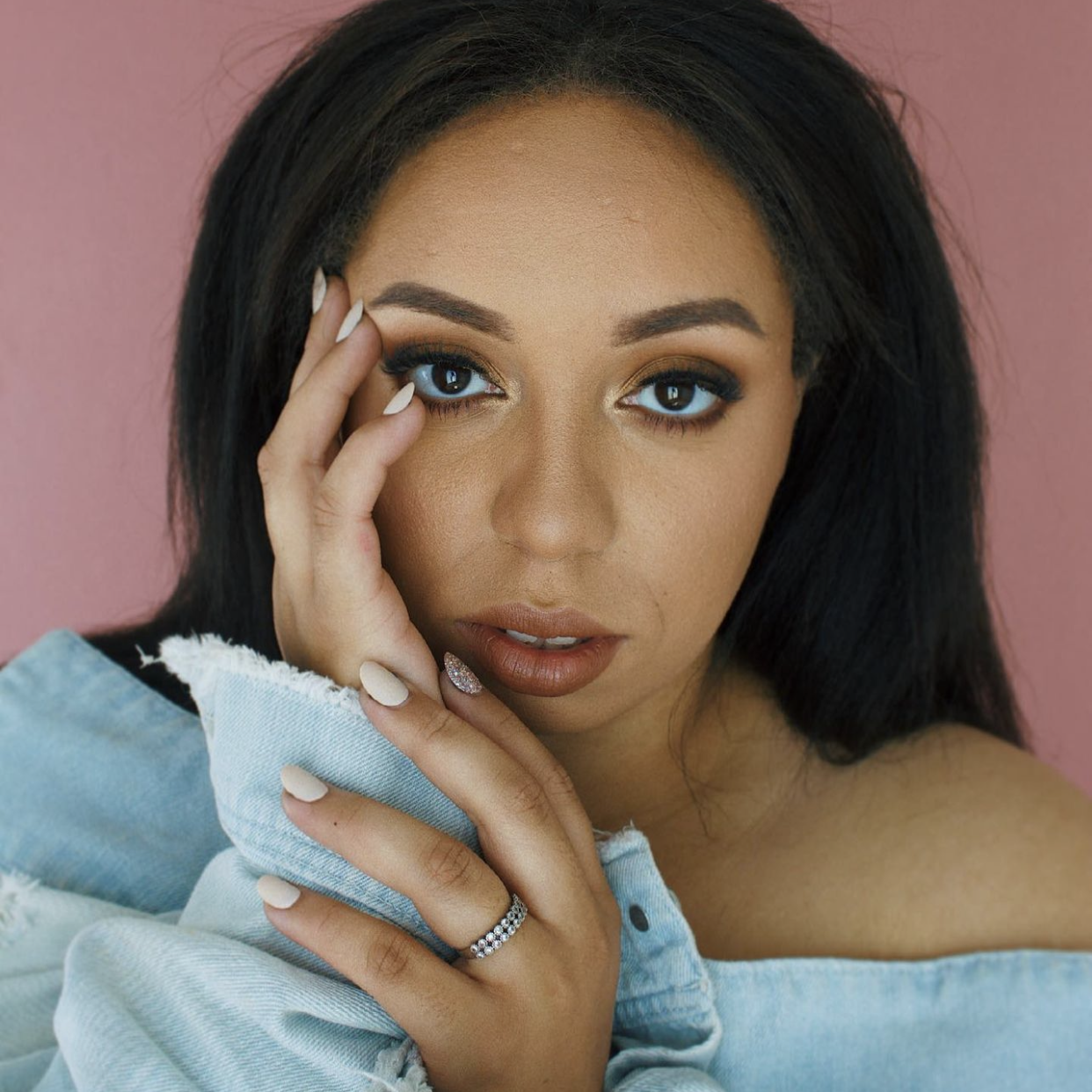 Close-up portrait of a woman with dark hair, blue eyes, and makeup, touching her face with her hand, wearing a light blue denim jacket and a silver ring, with a pink background.